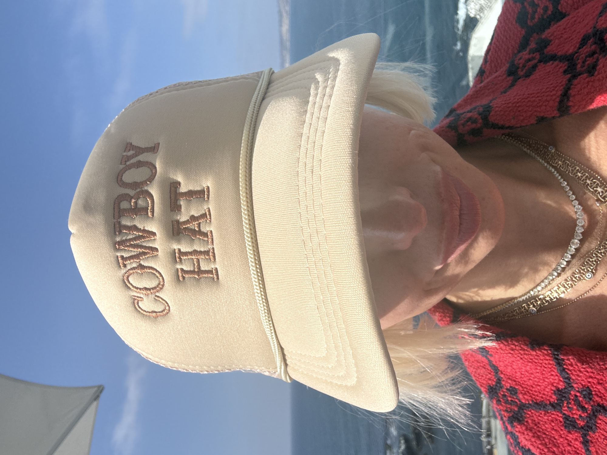 The image shows a person wearing a light-colored baseball cap that reads "COWBOY HAT" in embroidered text. The hat is positioned on the person's head at an angle, partially obscuring their face. The person appears to be wearing a red patterned sweater or top and multiple gold necklaces. The background consists of a body of water with a slight ripple, a blue sky, and what seems to be a partially visible white sail or canopy. Without further information, the location in San Diego cannot be determined.