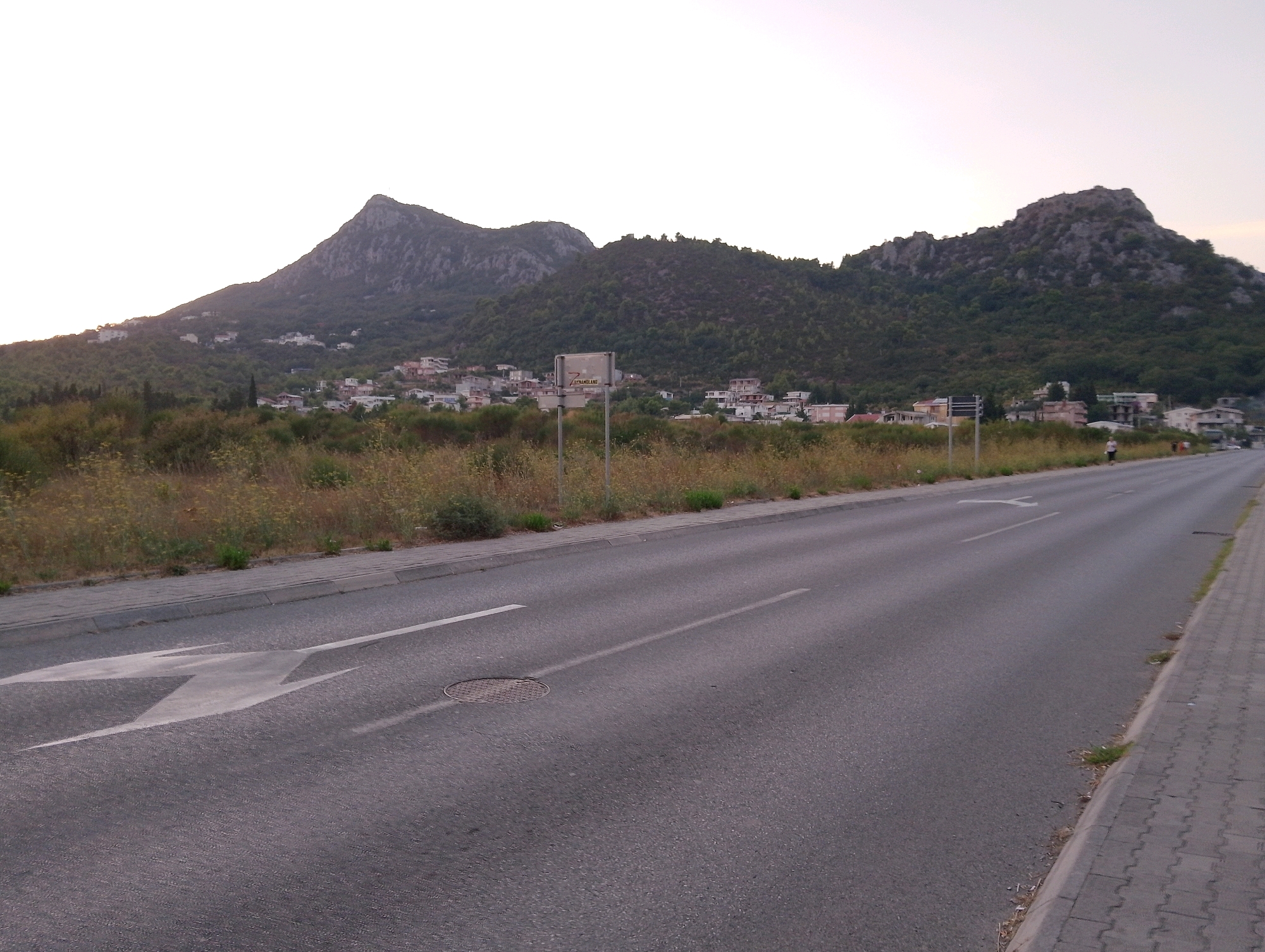 The image depicts a wide asphalt road in Sutomore, Montenegro, with a cobblestone sidewalk on the right. The road is marked with white lines, including a directional arrow painted on the asphalt. In the background, a town is nestled against lush green hills and rocky mountains, with scattered houses and buildings visible. The sky is overcast and light, suggesting either early morning or late evening. A single person is walking on the sidewalk in the distance. The scene is static and appears to be a typical street view in a coastal town.