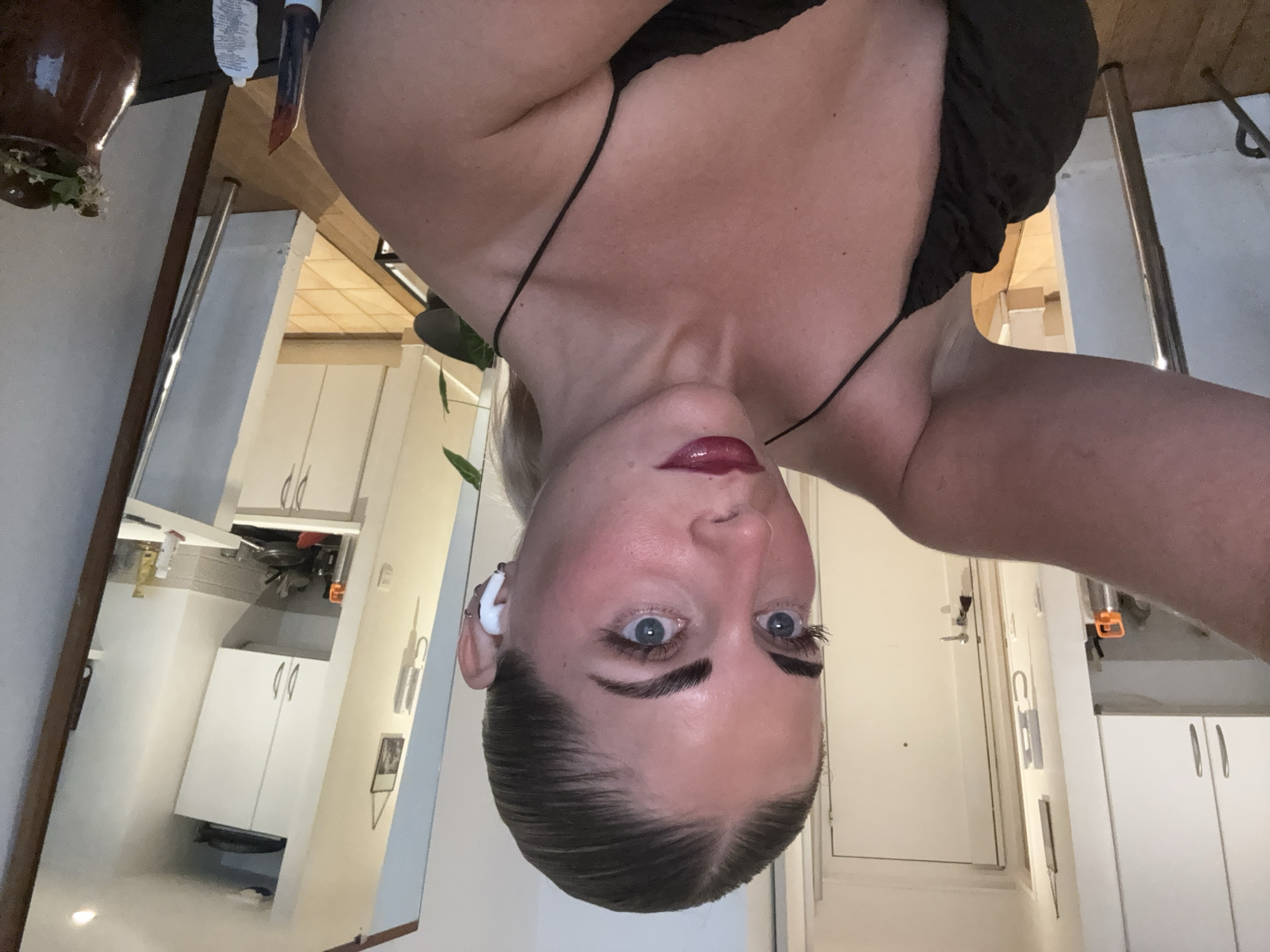 A woman is taking a mirror selfie in an apartment. She is wearing a black slip dress and has her hair pulled back in a ponytail. She is also wearing white earbuds. The apartment appears to be modern, with white cabinets and tiled floors visible in the background. A doorway is visible in the background, as well as a large mirror that reflects a portion of the kitchen.