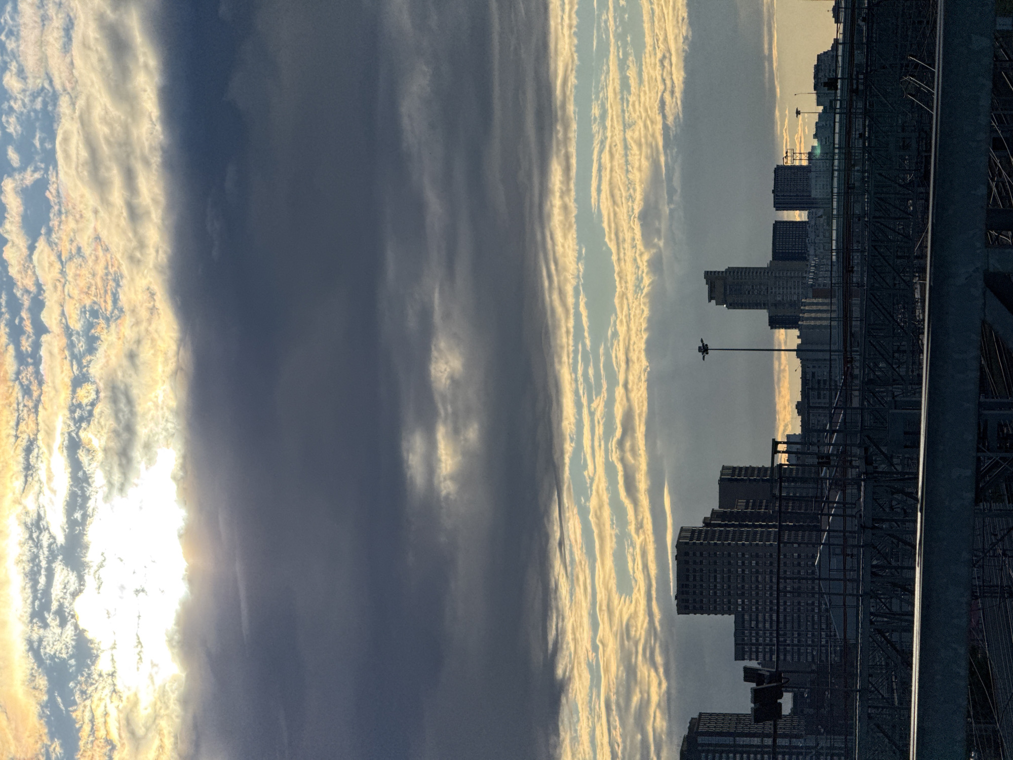 This media file depicts a cityscape with a dramatic sky. The primary subjects are a collection of tall buildings that form a city skyline, silhouetted against a sky filled with dynamic clouds. The clouds are illuminated by the sun, creating a contrast between areas of bright light and deep shadow. The sun itself is partially obscured by clouds, suggesting it is either rising or setting, casting a warm, golden hue on the edges of the clouds on the right side of the image.
The scene is set in a urban environment, as indicated by the dense concentration of high-rise buildings and the presence of what appears to be infrastructure such as elevated tracks or bridges, and streetlights, on the right side of the frame. The overall impression is one of a vast, metropolitan area bathed in the atmospheric light of either dawn or dusk.
Notable details include the texture and form of the clouds, which are wispy and elongated in some areas, and more dense and textured in others. The light suggests a time of day when the sun is low on the horizon, creating long shadows and highlighting the contours of the clouds. The weather appears to be mixed, with both bright, sunny patches and darker, cloud-covered areas. The location context of Moscow, Russia, suggests a potentially cold climate, though this is not explicitly visible. No people are clearly visible, but the scene implies human habitation and activity through the presence of the city. No text is visible in the image. The emotions evoked could range from awe at the natural beauty of the sky to a sense of the grandeur and scale of urban life.