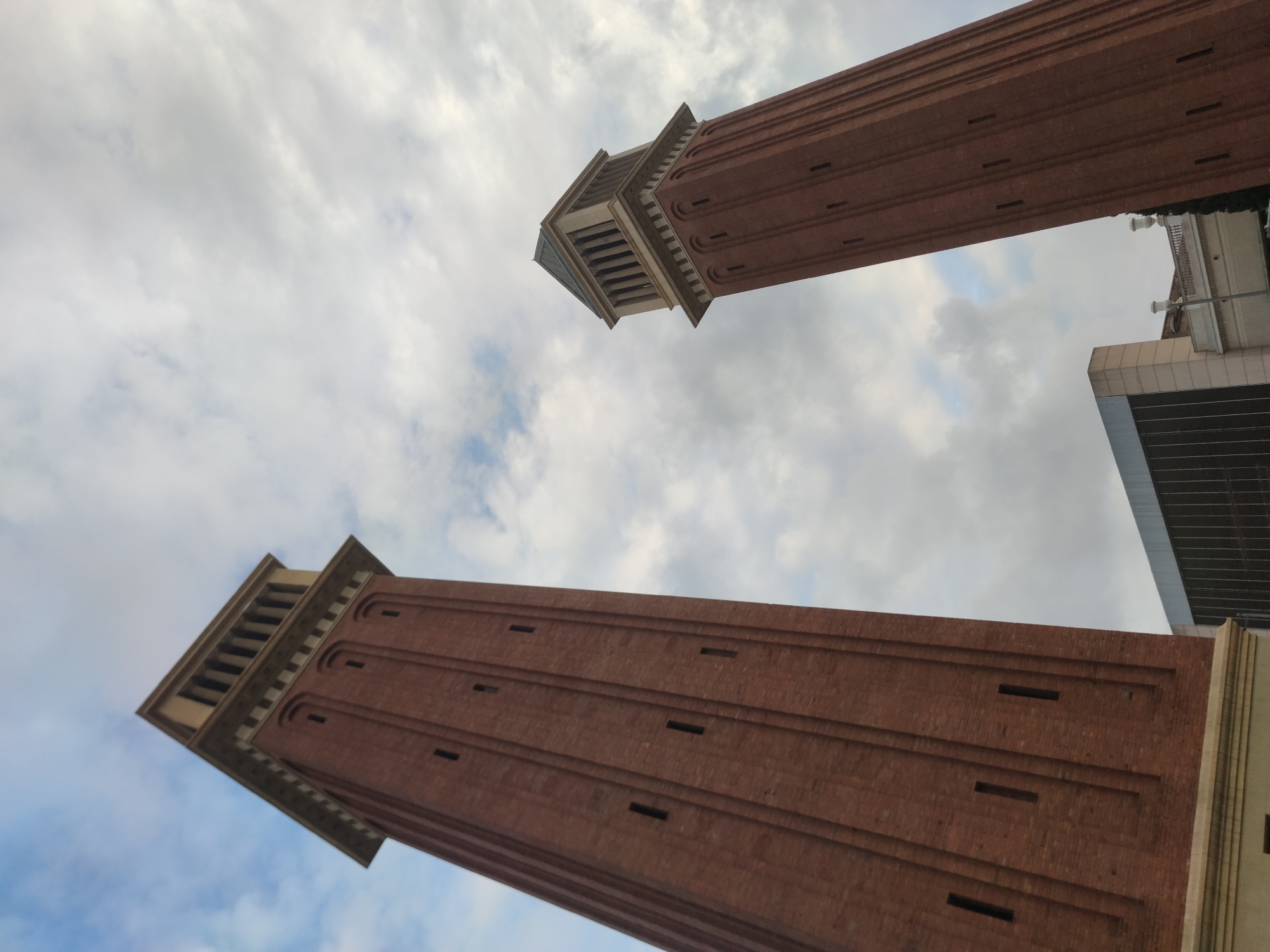 The image captures a low-angle view of the two Venetian Towers in Barcelona, Spain. The towers, constructed of red brick, stand prominently against a backdrop of cloudy sky. Each tower has a distinct, tiered top section, different from the tower bodies. A portion of a modern building with glass facade is visible on the right.