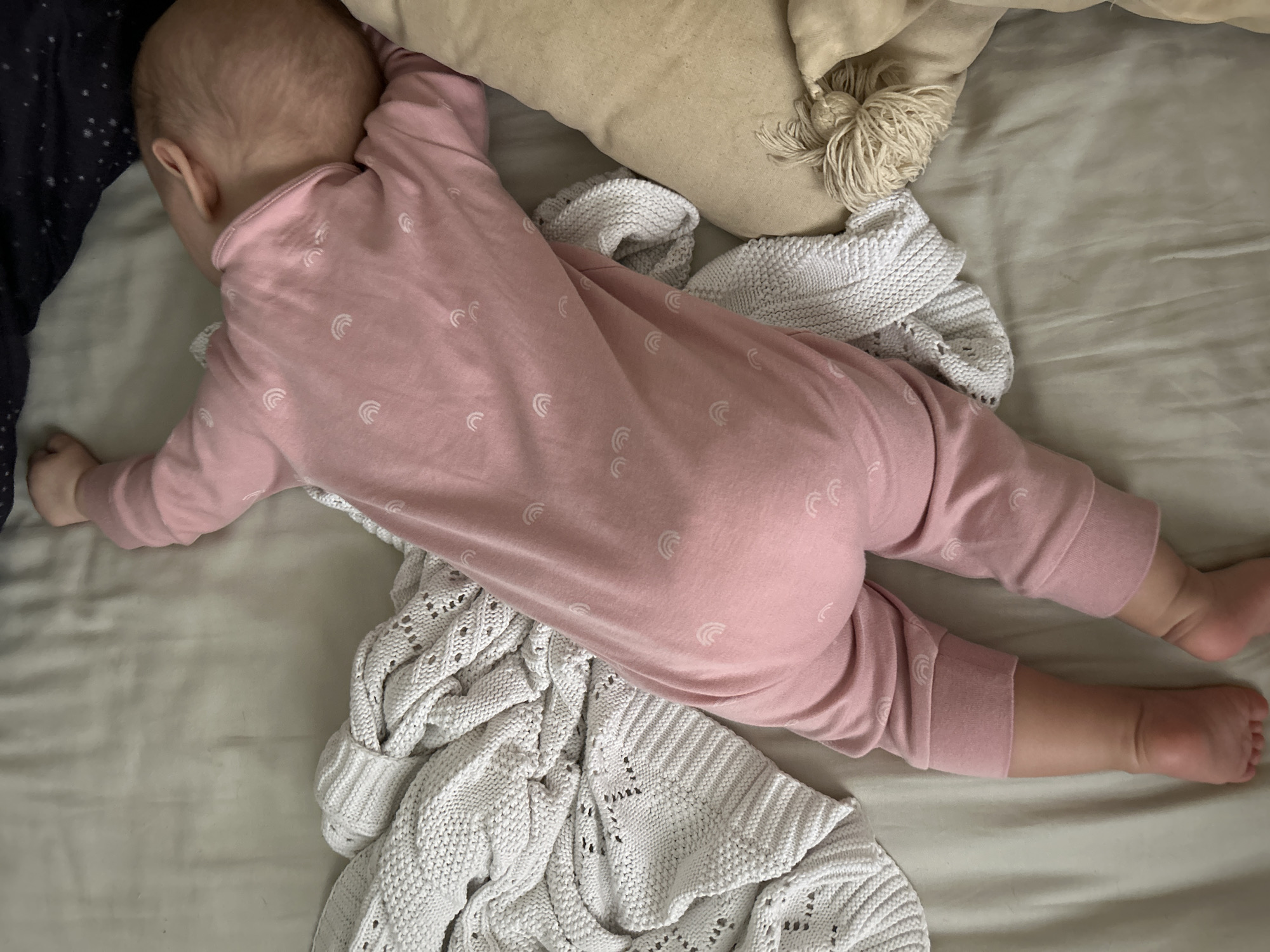 An overhead, close-up shot depicts a baby lying on its stomach, seemingly asleep or resting. The baby is wearing light pink pajamas adorned with small, white, crescent-shaped rainbow patterns. Its head is turned slightly to the left, partially obscured by a dark-colored fabric, possibly a blanket or sheet, visible on the far left of the frame.
The baby's body extends across a light, possibly cream or off-white, textured bed sheet. Beneath its torso and to its right, a white or cream-colored knitted or crocheted blanket with a distinct pattern of stitches and small eyelets is crumpled. Above the baby's head and upper back, a beige or light brown pillow with decorative tassels on its right side is visible. The lighting is soft and diffused, suggesting an indoor setting without harsh direct light. No specific location cues for Moscow, Russia, are visible within the image, nor is any text. The overall scene conveys a sense of calm and peaceful rest.