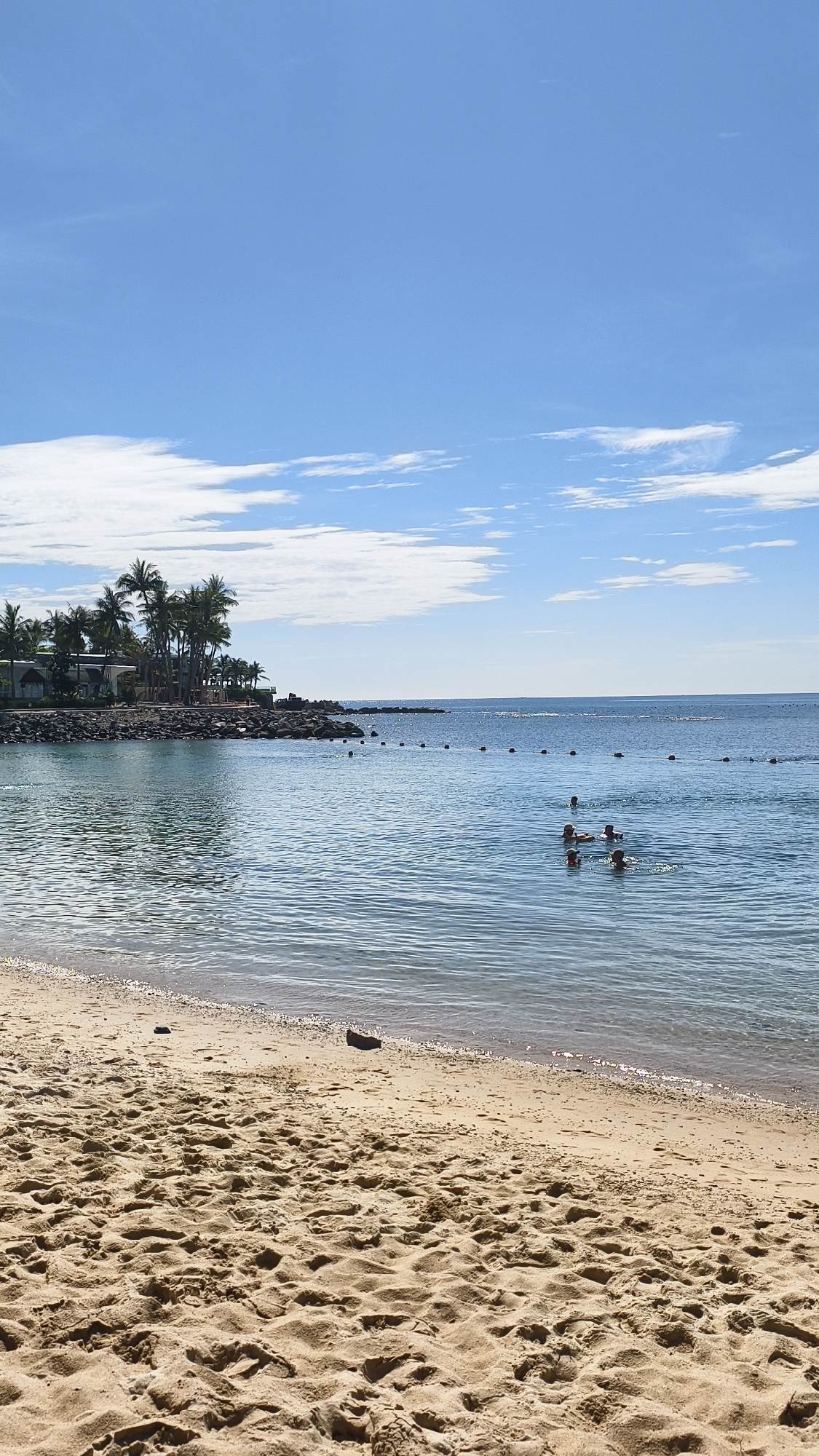 The image shows a beach scene in Nha Trang, Vietnam. In the foreground, there is a sandy beach with footprints. The middle ground features the ocean with several people swimming or floating on inflatable devices. A line of buoys marks a boundary in the water. In the background, there is a rocky shoreline with palm trees and buildings. The sky is blue with scattered white clouds.