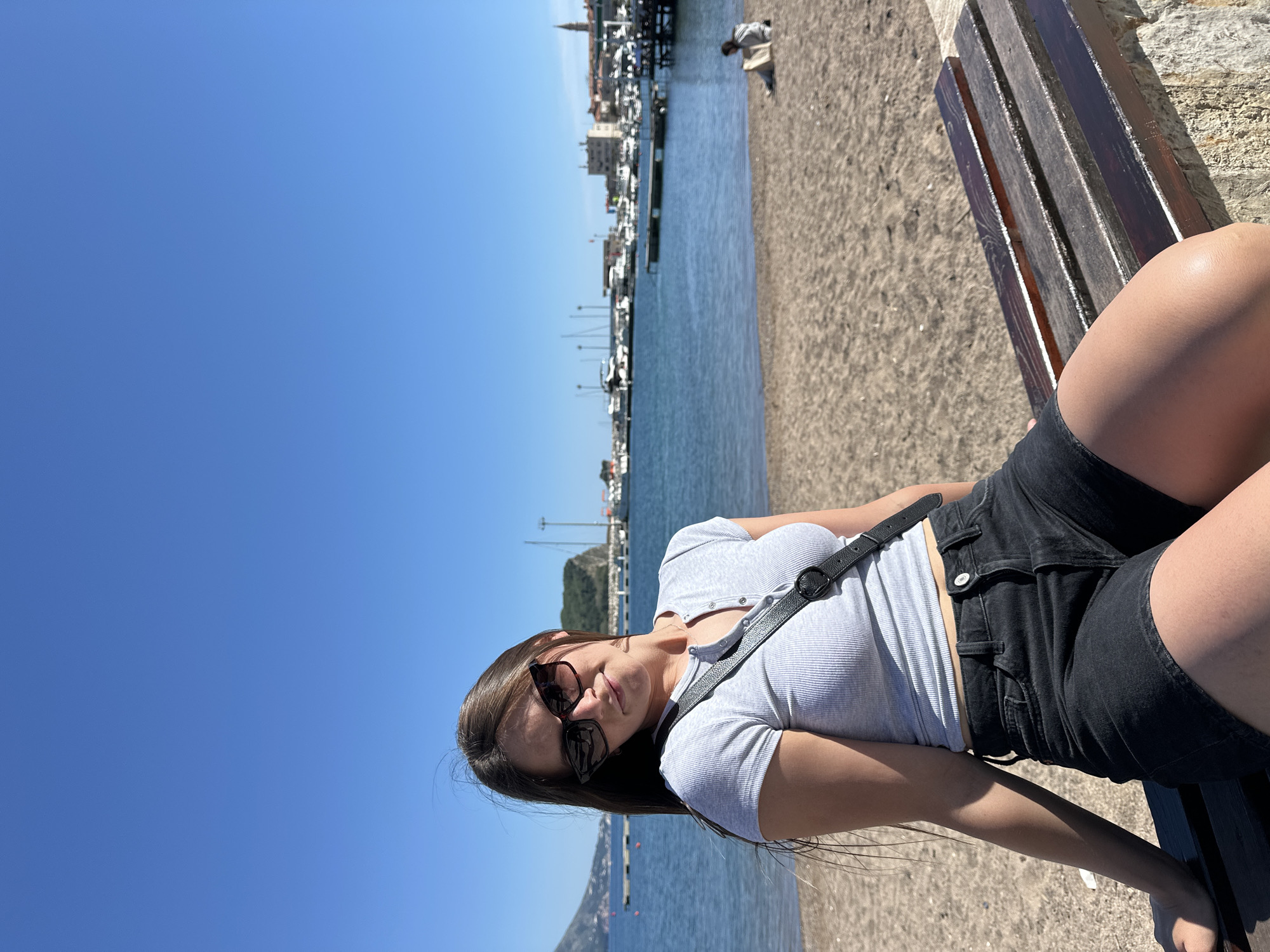A young woman wearing sunglasses, a gray ribbed short-sleeve shirt, and black shorts is reclining on a dark wooden bench. She has long brown hair. A black strap crosses her chest. The bench appears to be located on a sandy beach. Behind her is a body of water, and in the background, there is a cityscape with buildings and boats docked in a harbor. The sky is clear and blue. The location is most likely in the city of Budva, Montenegro, given the architectural style and coastal setting.