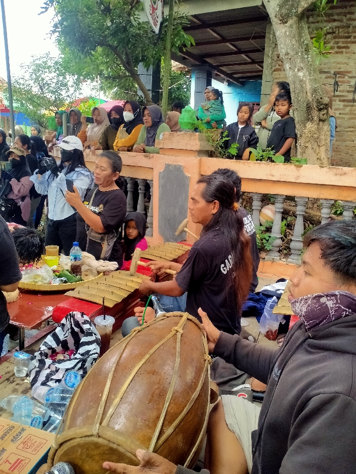 The image depicts an outdoor cultural gathering in Jenggrik, Indonesia. In the foreground, two men are performing traditional music. On the right, a man in a black hoodie and patterned neck gaiter plays a large, brown wooden kendang drum with his hands. Behind him, another man with long dark hair, wearing a black t-shirt, is seated and plays a gender, a metallophone instrument with a red frame and wooden keys, using two mallets. A microphone is positioned near the kendang drum. To the left of the musicians, on a small red table, a round woven tray contains food items, surrounded by plastic water bottles and cups with straws.
A crowd of onlookers, primarily women wearing headscarves and children, observes the performance from behind a low, light-colored concrete barrier with black and gray accents, and decorative baluster-like elements. Several individuals in the audience are actively recording the event with smartphones and at least one camera held by a woman in a light blue shirt and white cap. In the background, portions of a building with a corrugated metal roof and brick walls are visible, along with green foliage and tree trunks. A white and red emblem is affixed to a pole in the upper left section of the image.