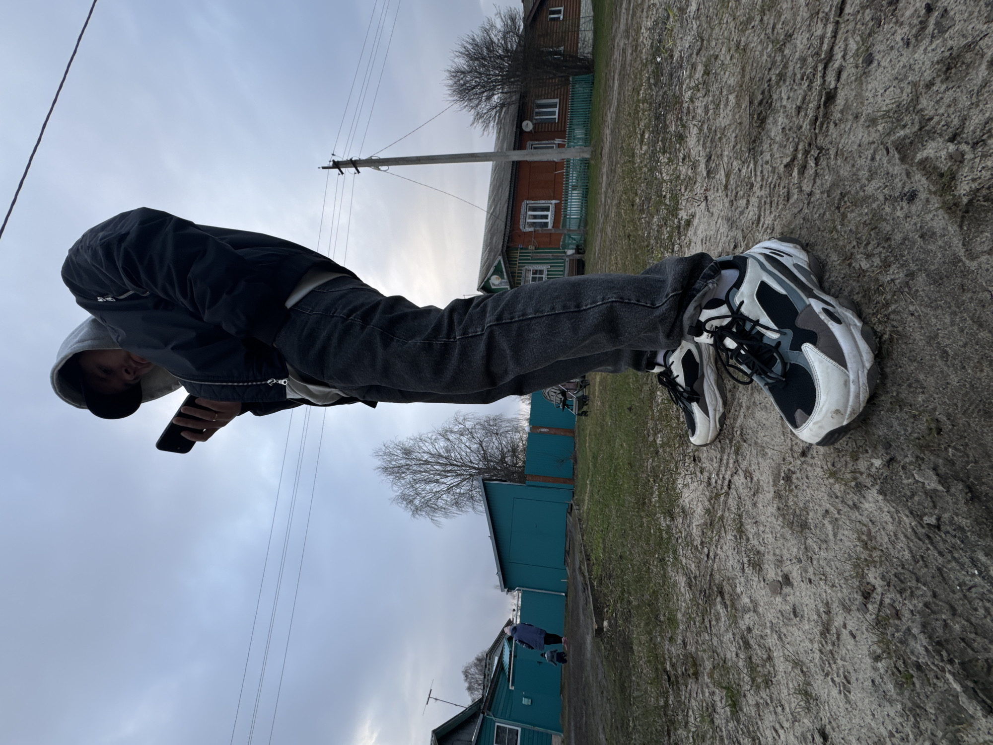 A low-angle photograph depicts a person standing in an unpaved area, likely in the city of Красный Маяк, Russia, based on the architectural style of the buildings in the background. The person is wearing black jeans, a black jacket, and sneakers with a black and white pattern. The person is holding a smartphone, presumably taking the photograph. The environment includes a dirt or gravel path, power lines, and a wooden house with green trim and red walls in the background, along with a row of turquoise-colored buildings. The sky is overcast.