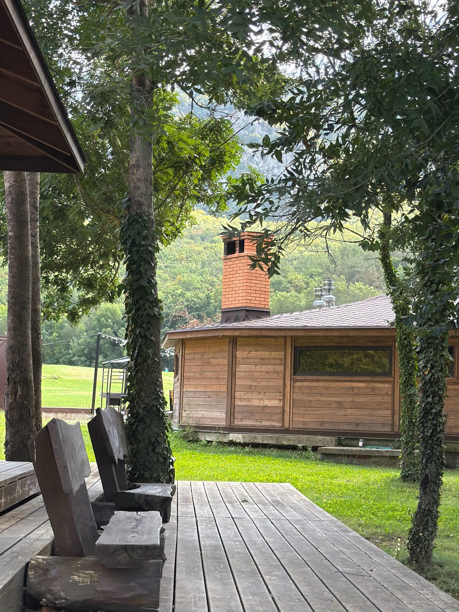 The image shows a wooden deck with rustic log benches in the foreground, overlooking a grassy area with a wooden building in the background. The building has a dark roof with a brick chimney and a long, narrow window. Trees with lush green foliage surround the scene, with a gently sloping, tree-covered hill visible in the distance. The overall impression is of a peaceful, natural setting.
