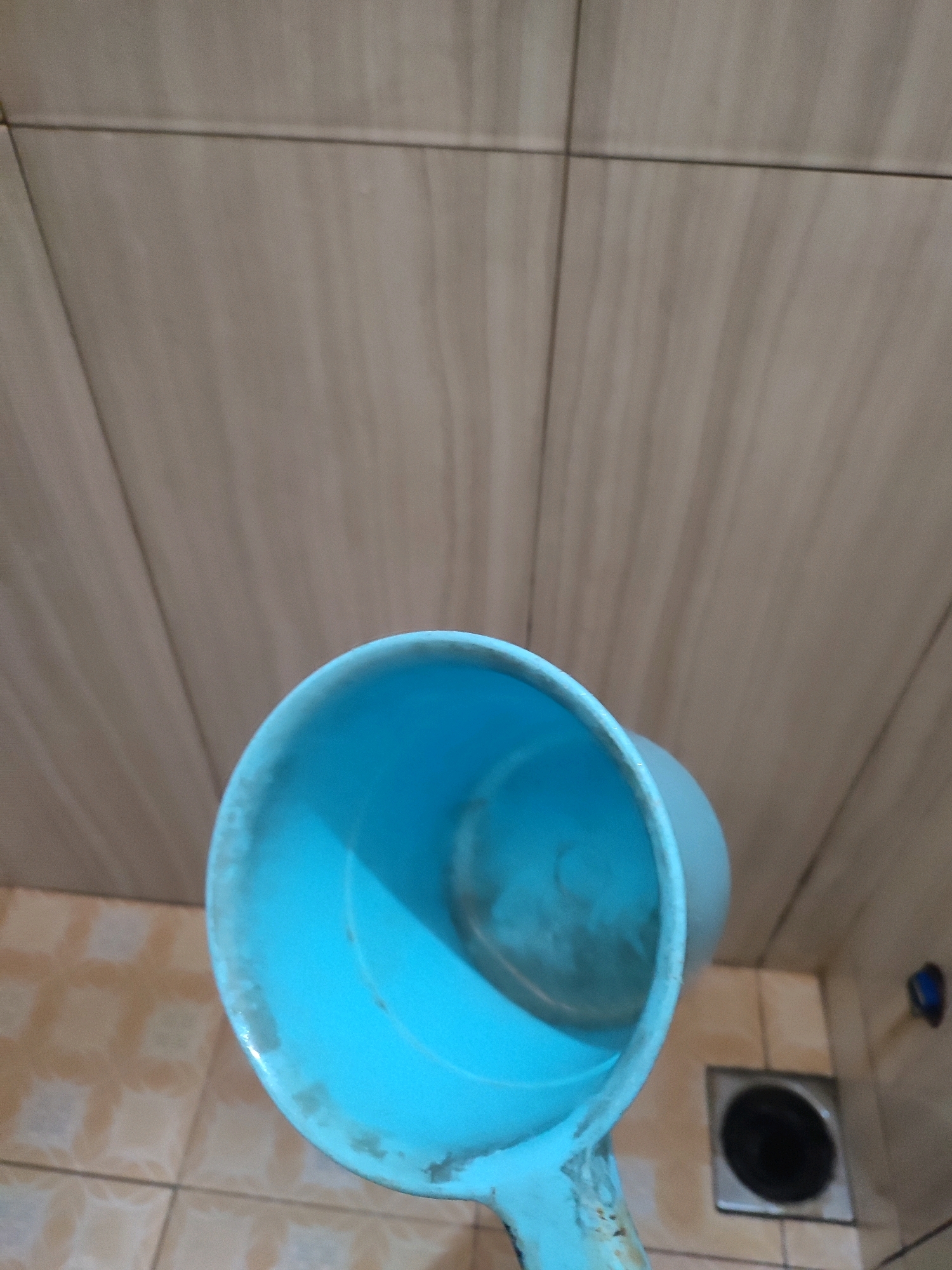 An upward-angle shot captures a light blue plastic dipper or ladle held in a bathroom. The dipper, showing some wear and discoloration, is positioned in the foreground. In the background, light beige tiles cover the wall and floor, with a dark circular drain grate visible on the floor. A dark blue object, possibly a faucet or handle, is mounted on the wall to the right of the drain. The scene appears to be in a bathroom, likely in a private residence in Bandung, Indonesia, given the typical tiling and fixtures common in the region. No people are present in the image.