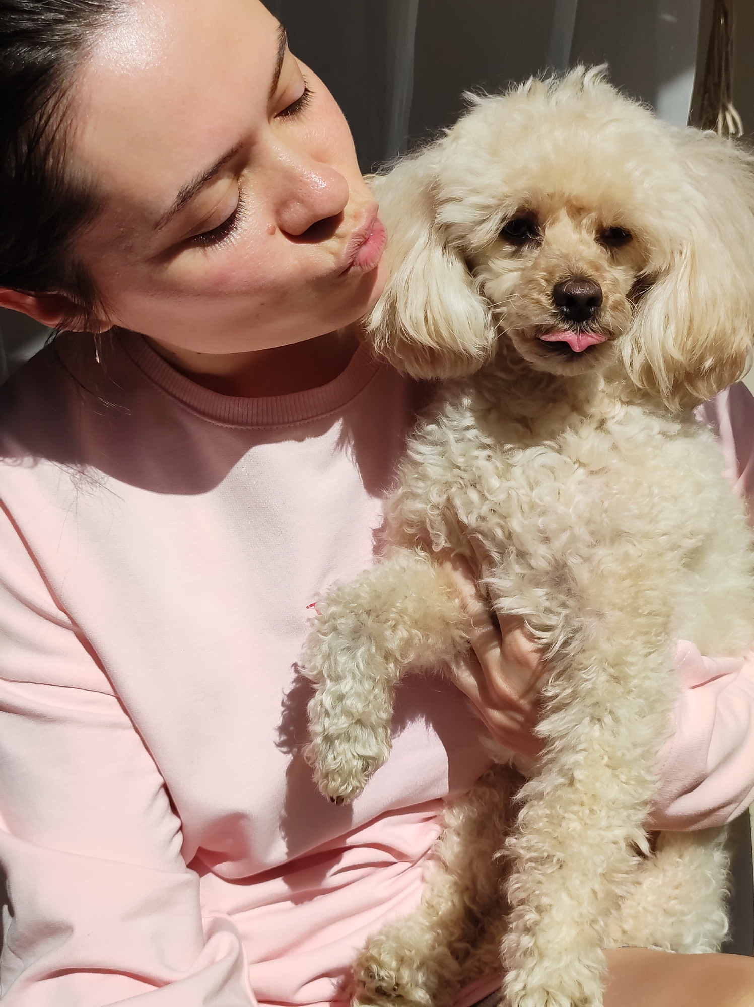 A young woman in a pink long-sleeved shirt holds a cream-colored poodle in her arms and puckers her lips in a kissing gesture towards the dog. The dog sticks its tongue out. The scene is illuminated by sunlight, casting shadows across the woman's shirt. The location, Chaikovsky, Russia, cannot be determined from the visual data.