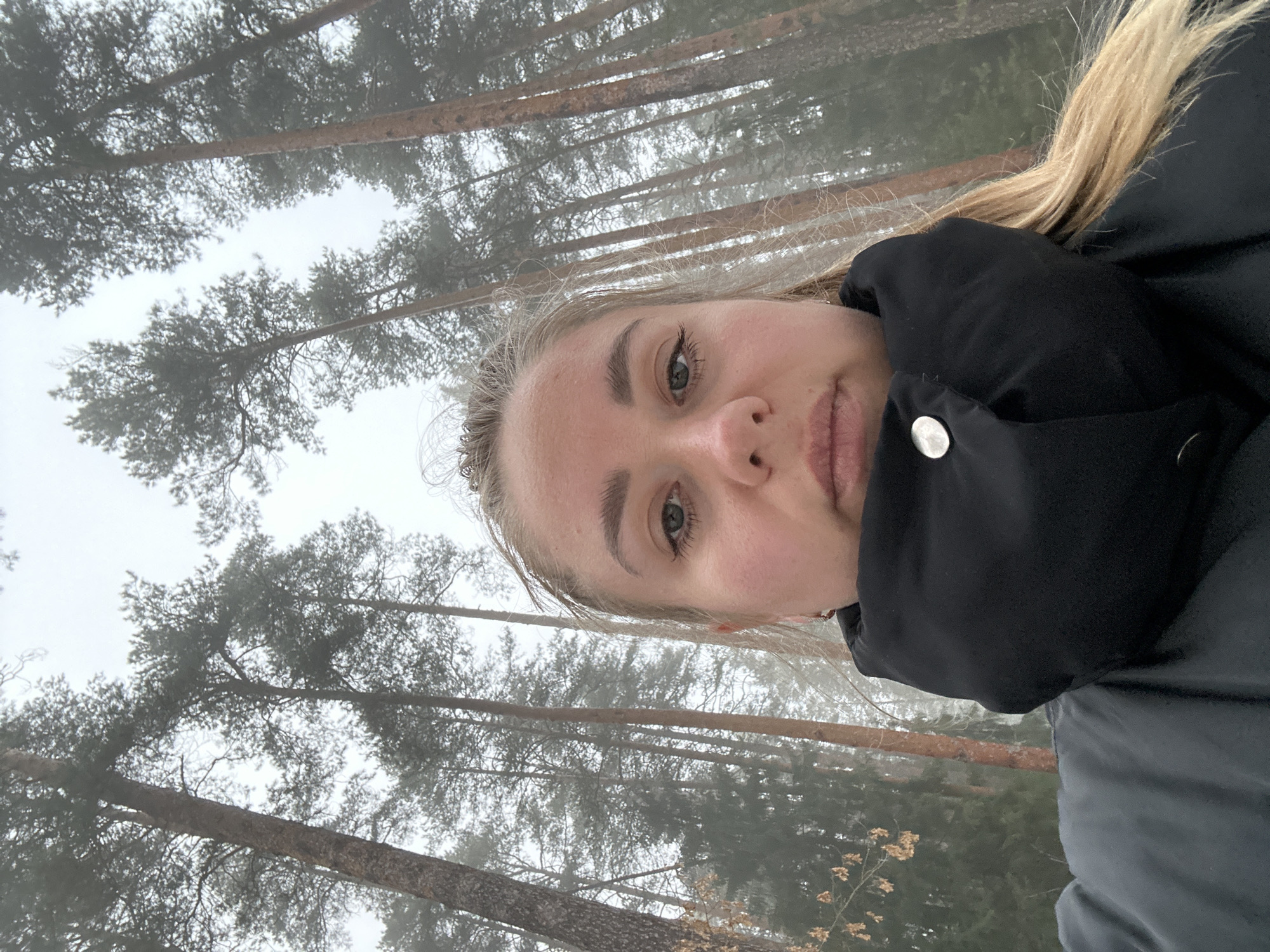 The image shows a selfie of a blonde-haired woman in a black jacket with a silver button, taken in a wooded area. The trees are tall and thin, with branches reaching into a foggy sky. The woman is looking at the camera with a neutral expression. The background appears to be in the city of Chaikovsky, Russia, based on the type of trees and the foggy weather, but there is no specific location information available from the image.