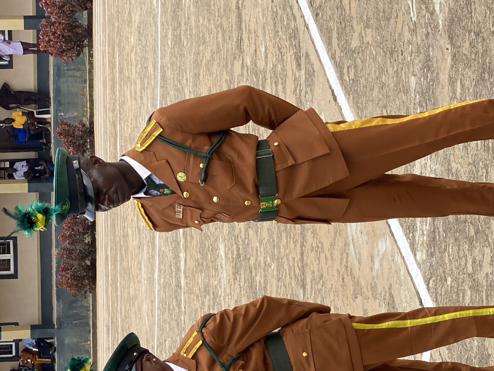 The image captures a formal outdoor setting, likely a parade ground or ceremonial area, in Kaduna, Nigeria. Two individuals in military-style brown uniforms with green and gold accents are the primary subjects. The man in the foreground is wearing a peaked cap adorned with green and yellow feathers, a dark green tie with a crest, and a dark green belt with a silver buckle featuring a crest. His uniform includes gold epaulets and gold buttons. The second individual, to the left and partially visible, is also in a similar brown uniform. Behind them, a building with windows and a dark trim is visible. Several people are seated or standing in the background, observing the event. The ground appears to be a paved surface marked with white lines. The overall atmosphere suggests an official ceremony or gathering.