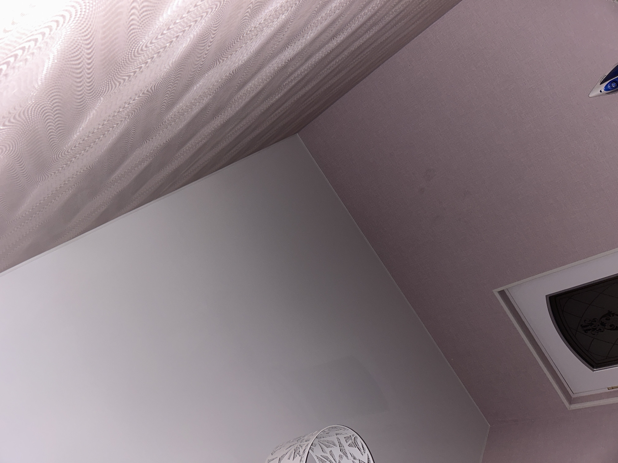 The image depicts an interior domestic space located in Charentsavan, Armenia. The scene is captured from an upward-angled perspective, showcasing a light-colored ceiling, walls, a portion of a door, a light fixture, curtains, and an iron.
The ceiling, occupying the upper left and central portion of the image, is a smooth, reflective, off-white or very pale grey surface, possibly a stretch ceiling, showing subtle, soft reflections. The walls are covered in a textured wallpaper of a uniform pale purple or dusty pink hue, displaying a fine, linen-like weave pattern.
In the upper left, a segment of a white, decorative lampshade is visible, featuring intricate geometric or fractal cut-out patterns. To the right, approximately one-third of the image width, light-colored sheer curtains or fabric are present, exhibiting a distinct wavy or rippled texture with subtle horizontal stripe patterns.
In the lower center-left, a white door frame encloses a dark-paneled door. The door's panel features a decorative pattern, possibly etched glass or an iron grille design. In the lower right corner, the front tip and a small section of a blue and white electric iron are visible, positioned against the pale purple wall.
No individuals are present in the scene, and no overt actions or interactions are occurring. The environment appears clean and well-maintained, illuminated by an unseen light source that casts soft, even lighting across the surfaces.