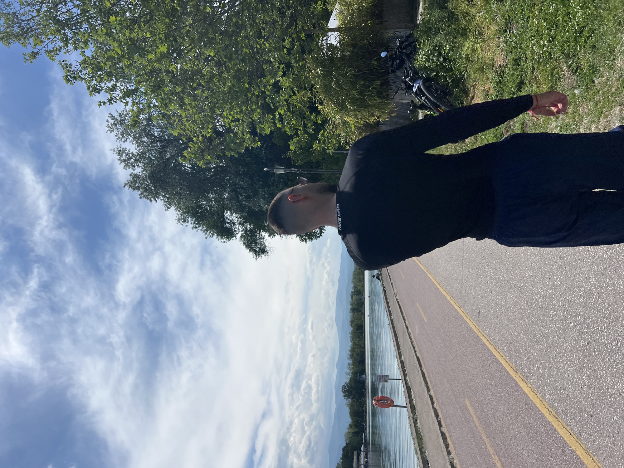 A man is walking along a paved path next to a body of water. The water extends to the horizon, where there are mountains. The sky is blue with clouds. A tree and foliage are to the left of the man. The man is wearing dark clothing. A bicycle is parked near the tree. An orange life preserver is visible near the water's edge. The location is on the Maritza River bike path in Plovdiv, Bulgaria.