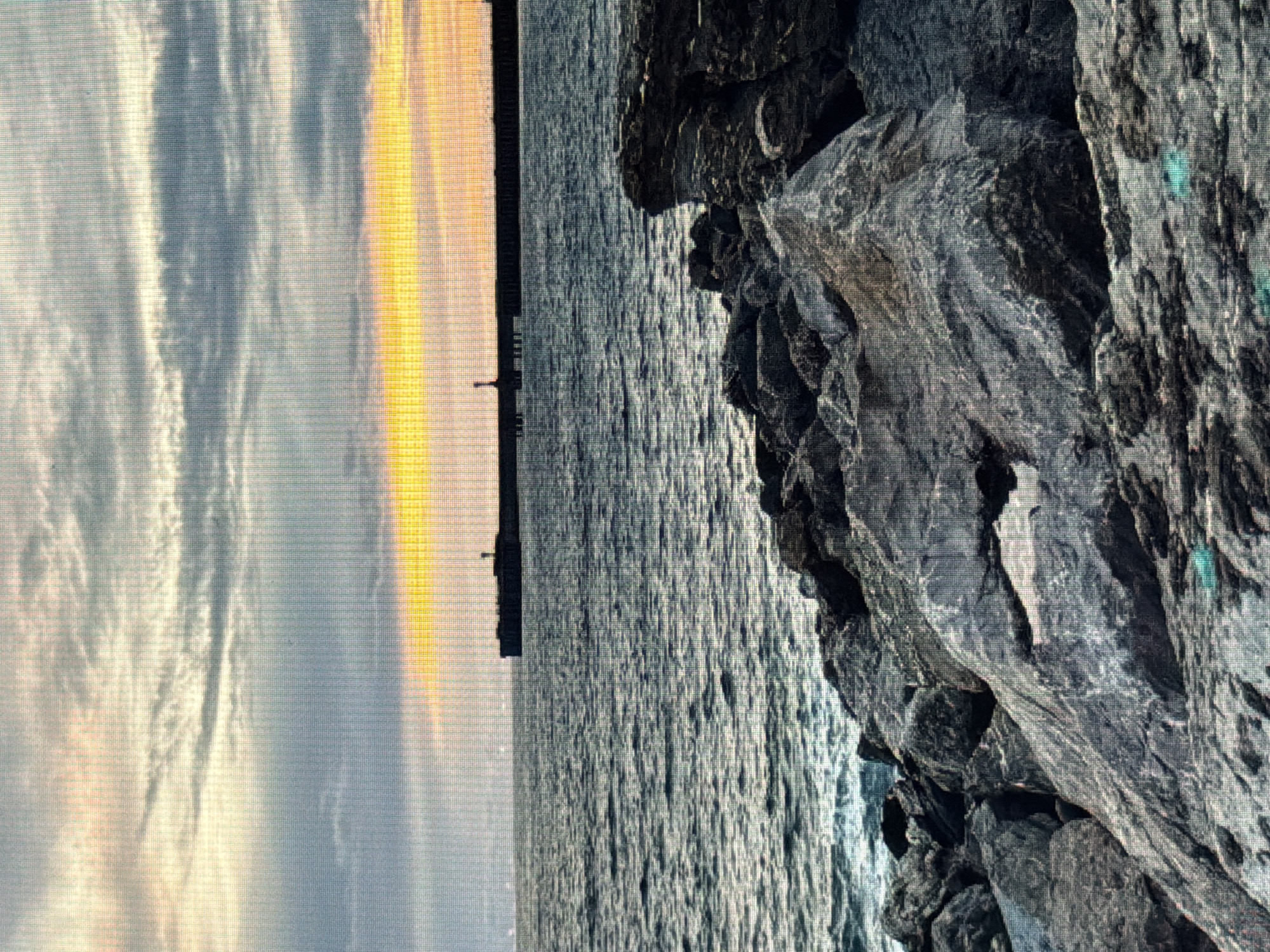 The image depicts a rocky shoreline with the ocean in the background. A long, dark structure, possibly a pier or breakwater, extends across the horizon. A bright band of yellow and orange light, indicative of a sunset or sunrise, is visible in the sky above the horizon. The water is a dark blue-gray, with visible waves. The foreground is dominated by large, gray rocks, some of which appear rough and weathered. The sky is cloudy, with streaks of gray and lighter tones. There are no people or animals visible in the image. The scene appears to be a natural coastal landscape. The location of Sirius, Russia, is not visually identifiable within this image.