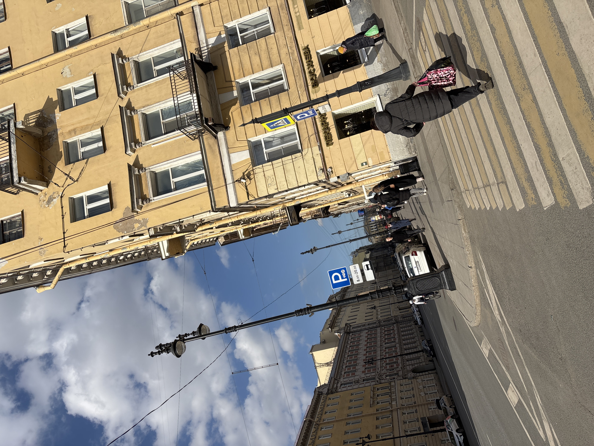This image depicts a sunny street scene at the intersection of Gorokhovaya Street and Bolshaya Morskaya Street in Saint Petersburg, Russia.
The foreground features a wide asphalt road with a pedestrian crosswalk marked by white and faded yellow stripes. A person, viewed from behind, wearing a dark puffer jacket and dark pants, is walking diagonally across this crosswalk from left to right, carrying a pink and black floral-patterned tote bag.
To the right, a multi-story yellow building with classical architectural features, white-framed windows, and several black-railed balconies dominates the scene. Its facade shows signs of wear, including peeling paint and discoloration. The ground floor houses retail establishments, one with a sign reading "ПЕКАРНЯ" (Bakery) and another "ВКУС ЖИЗНИ" (Taste of Life). A tall, ornate black streetlight pole with multiple luminaires stands prominently near this building, bearing a blue square parking sign ("P 8.00-20.00") and a yellow and blue pedestrian crossing sign. Another pedestrian in a yellow hat and dark coat, carrying a green bag, is visible on the sidewalk in front of these shops.
To the left, a row of similar multi-story buildings, mostly yellow, lines a wide street stretching into the distance. Several cars are parked along this street. A person on a bicycle, wearing a white top, is visible in the roadway further down the street. Additional pedestrians are visible on the sidewalks on both sides of the street.
The sky is bright blue with scattered white cumulus clouds. Overhead electrical wires are visible crossing the upper portion of the frame. Long shadows cast by buildings and individuals indicate the sun is at a relatively low angle.
