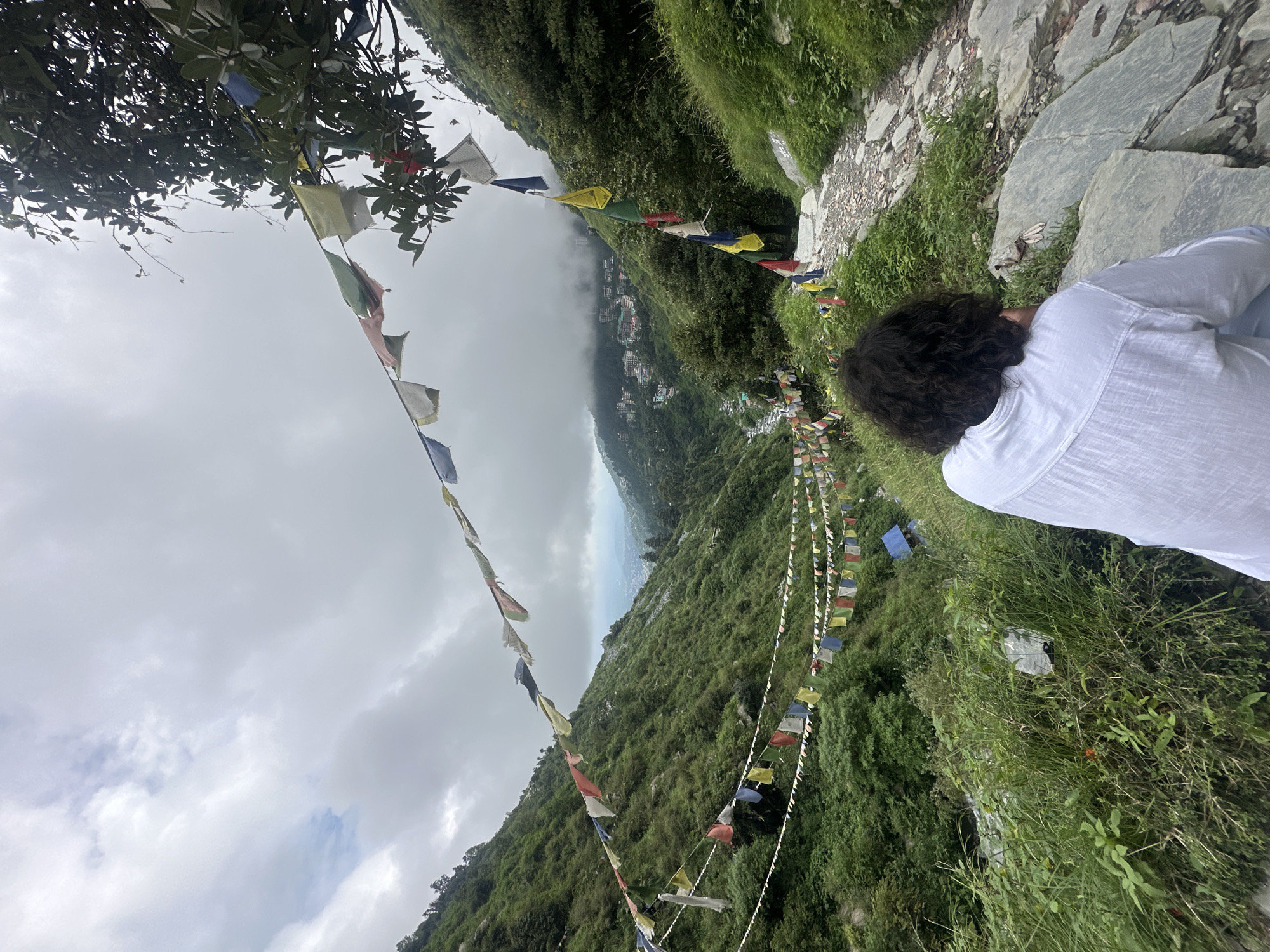 A lone individual, seen from behind, sits on a stone pathway overlooking a lush green valley. The person has dark, curly hair and wears a white shirt. Prayer flags, in various colors, are strung across the scene, some hanging from trees on the right and others stretched taut across the valley in the distance. The valley itself is densely forested, with a river or stream visible winding through it. In the far distance, a town or city is nestled among the hills, partially obscured by clouds. The sky is overcast with gray clouds. The pathway is made of rough stones and is bordered by green grass and foliage. The overall scene suggests a location in a mountainous region, likely Bhagsu, India, known for its natural beauty and spiritual sites.