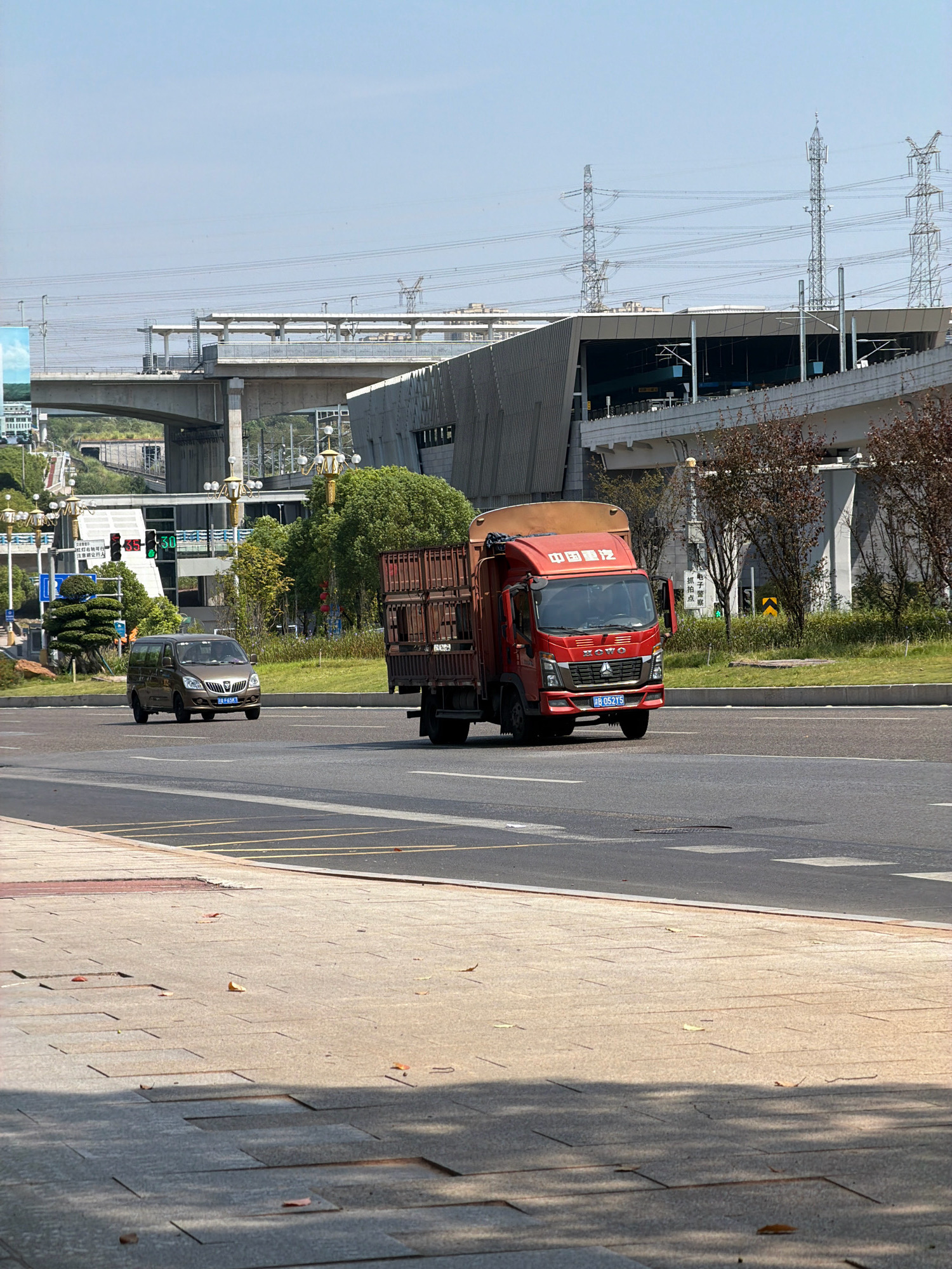 A red truck with a cargo of crates is driving on a city street in Jiangjin, China. A brown minivan is also driving on the street. In the background, there is a modern train station with overhead power lines and tall towers. The sky is clear and blue. The street is paved with asphalt and bordered by a sidewalk made of stone tiles. Some scattered leaves are on the sidewalk.