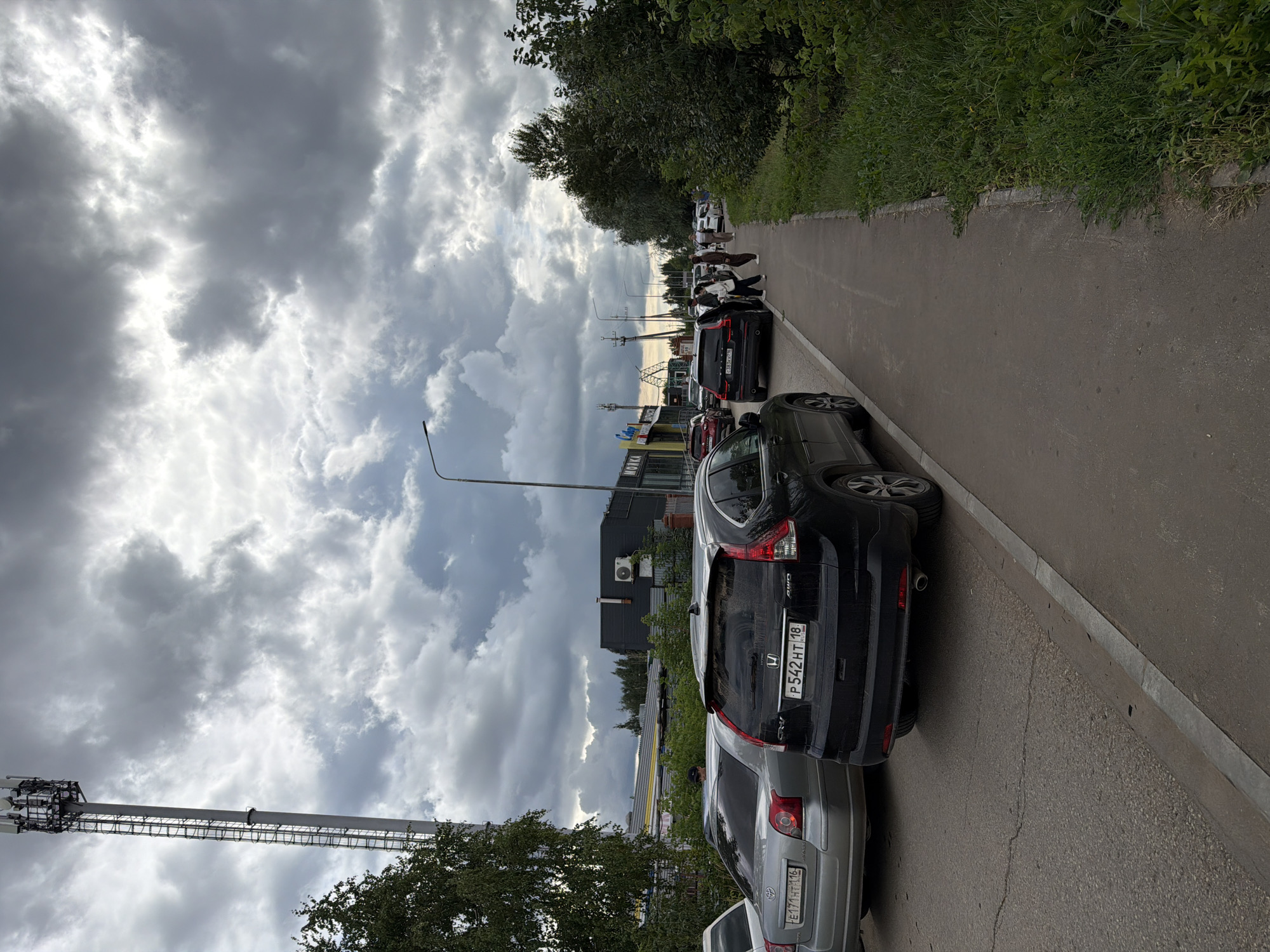 The image depicts a street scene in Yelabuga, Russia, with a prominent cell tower on the left. Several cars are parked along the road and on a sidewalk area. A black SUV, possibly a Honda CR-V, with license plate P542HT 18, is parked on the right side of the road. A silver sedan, a Toyota Avensis, with license plate E171HT 116, is parked to its left. Several people are visible, some standing near the vehicles and others walking along the sidewalk. The sky is heavily clouded, suggesting overcast weather. Buildings, including what appears to be a commercial establishment with a sign, are present in the background, along with trees and vegetation bordering the sidewalk. The ground surface is asphalt.
