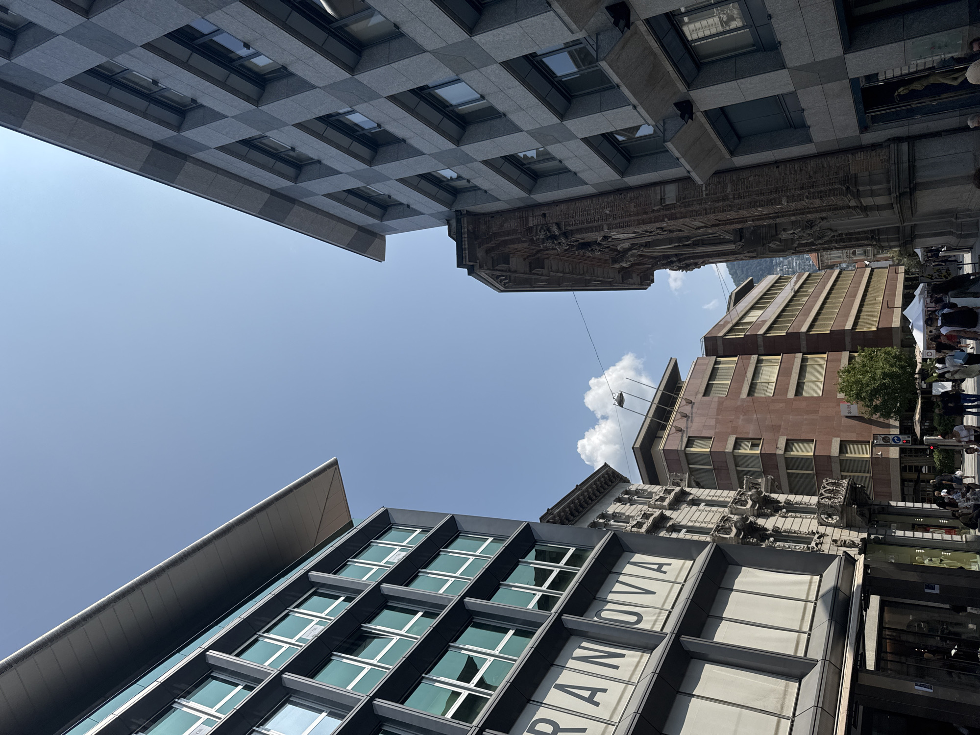 This image depicts a street scene in Lugano, Switzerland, under a clear blue sky with sparse clouds. The perspective is from street level, looking upwards at the facades of several buildings. On the left, a modern building with a dark gray exterior features large windows and the partially visible signage "GRANOVA". Adjacent to it, an older, ornate building made of brick with decorative stone carvings is visible. To the right of this, a grey, checkered facade building rises. Further down the street, another building with a reddish-brown facade and horizontal windows is present. The street is populated by a number of people walking and congregating, some near market stalls. A traffic light is visible on the right side of the street. The overall impression is a bustling urban environment with a mix of contemporary and historic architecture.