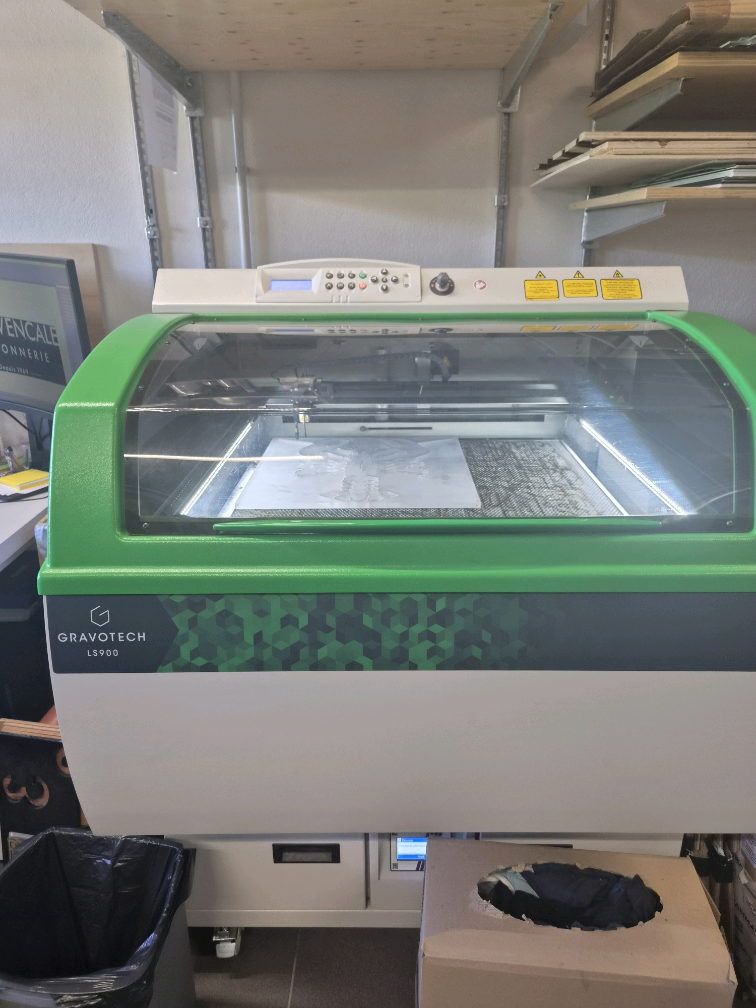 A Gravotech LS900 laser engraver is positioned in what appears to be a workshop or studio environment. The machine is predominantly white with a bright green hood that covers the laser apparatus and workspace. The hood has a transparent section allowing visibility into the engraving area, where a patterned material, possibly being cut or engraved, is visible. The Gravotech logo and model name are clearly displayed on the front of the machine.
Above the laser engraver, a metal shelving unit is attached to a pale wall, holding various flat items that appear to be sheets of material, possibly for engraving or printing. To the left of the engraver, a monitor displays a logo and text which includes "VEN CALE CONNERIE" and "Depuis 1969", suggesting a business or brand name.
In the foreground, a black trash bin is placed to the left of the machine. To the right, a large cardboard box with a roughly cut opening is visible, and it contains dark materials. Part of a roll of paper or similar material is also visible on the right side. The flooring is tiled. The scene is well-lit, indicating an indoor space. Based on the context, this workshop is located in Leudelange, Luxembourg, as indicated by the provided location information. There are no people visible in the image.