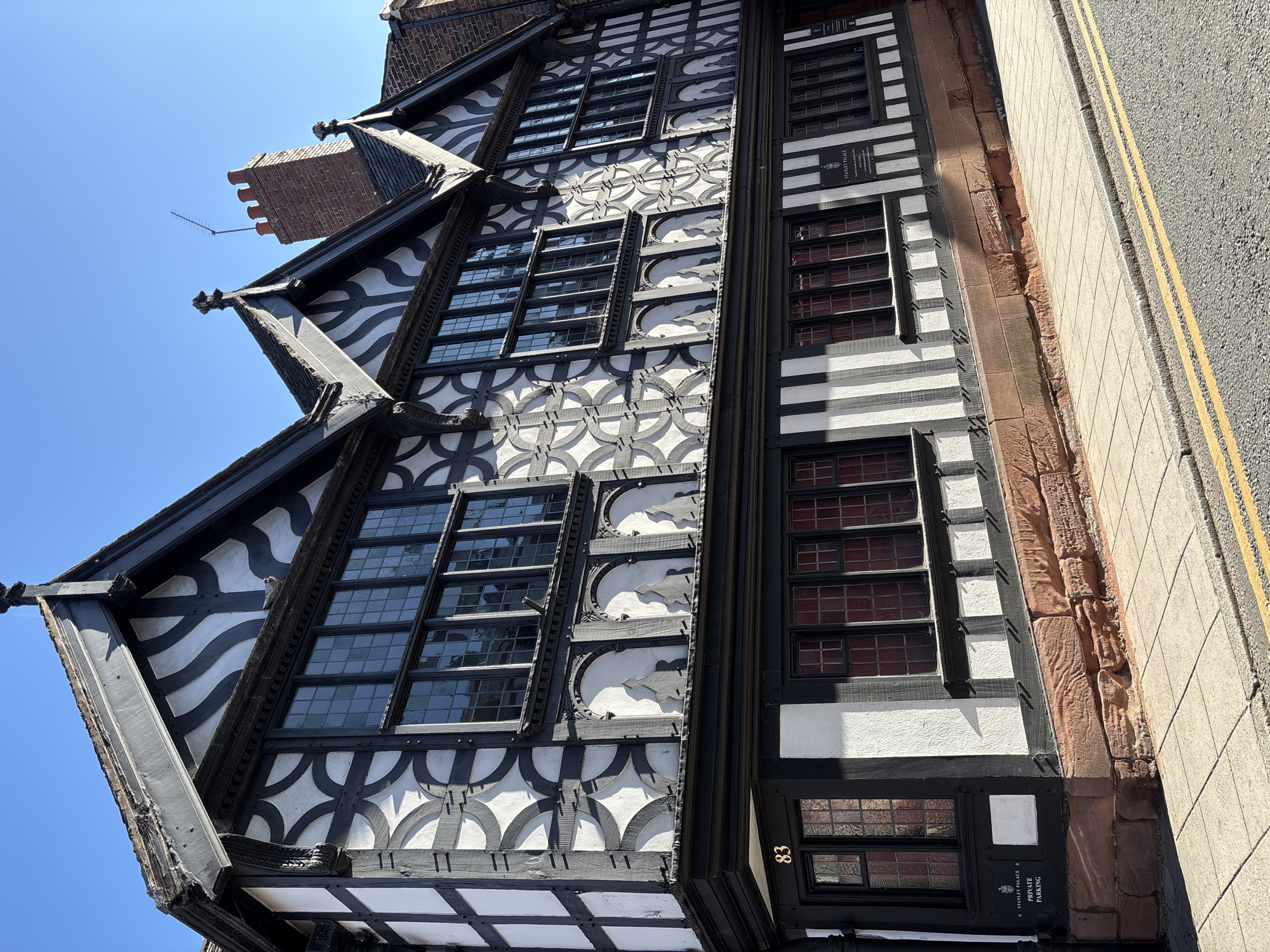 The image captures Stanley Palace in Chester, United Kingdom, a notable example of Tudor architecture. The three-story building features black and white timber framing, a hallmark of the period. The framing is adorned with decorative patterns. Multiple paned windows are present across all floors. A brick chimney rises from the roof. The side of the building abuts a sidewalk and a street with double yellow lines. A sign near the base of the building reads "Stanley Palace Private Parking" along with the building's number, 83. The sky is clear and blue. The image is rotated slightly, with the top of the building leaning towards the left.