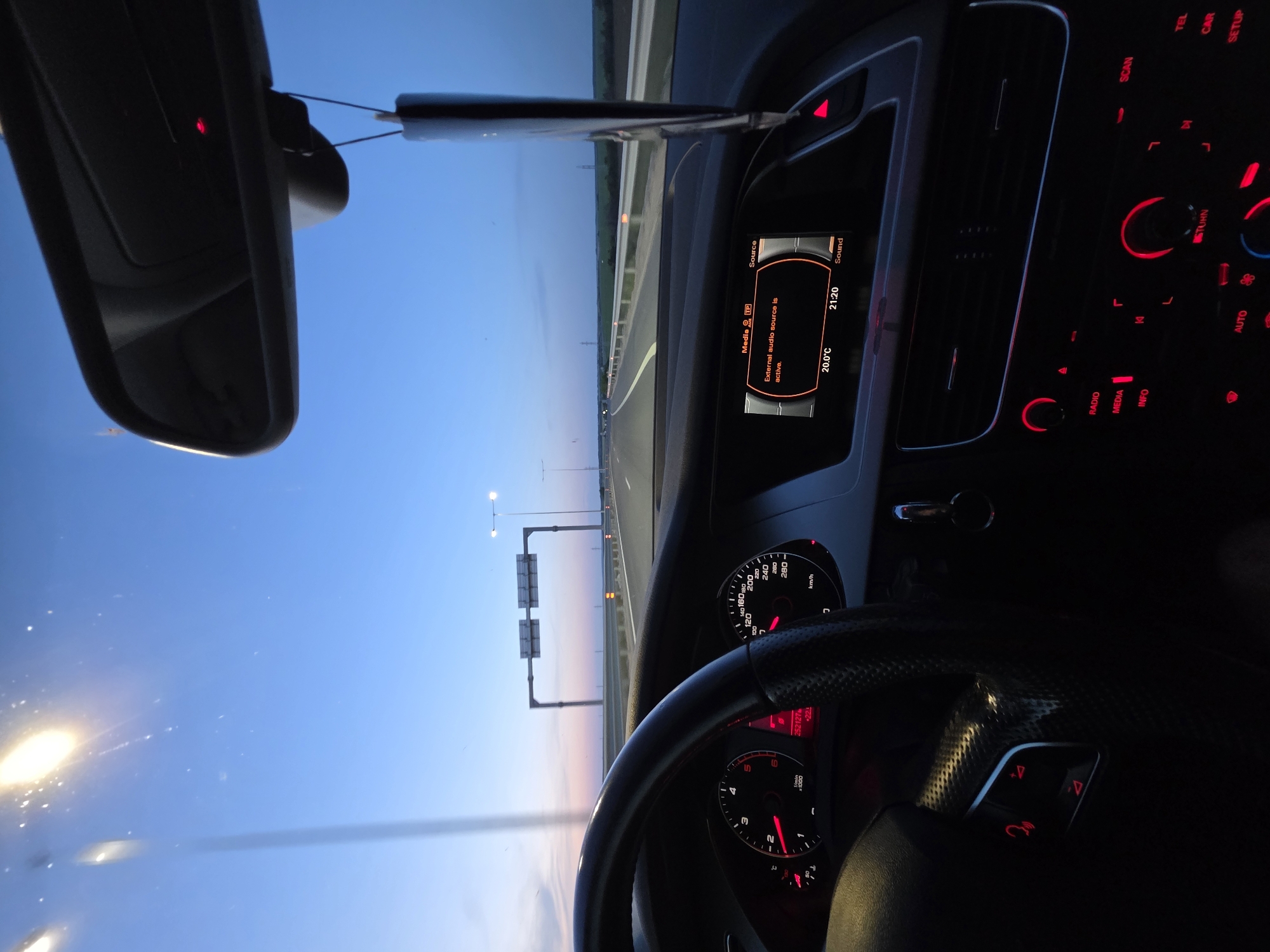 The image shows the interior of a car driving on a highway at dusk. The dashboard is visible, displaying information like speed (partially shown), engine RPM, and audio settings. The central console features illuminated buttons for radio, media, and climate control. The driver is in the car. Outside, the highway stretches into the distance with overhead signs and light poles. The sky is a gradient of colors indicating either sunrise or sunset.
It is impossible to tell if the image was taken in or near Dimovo, Bulgaria.