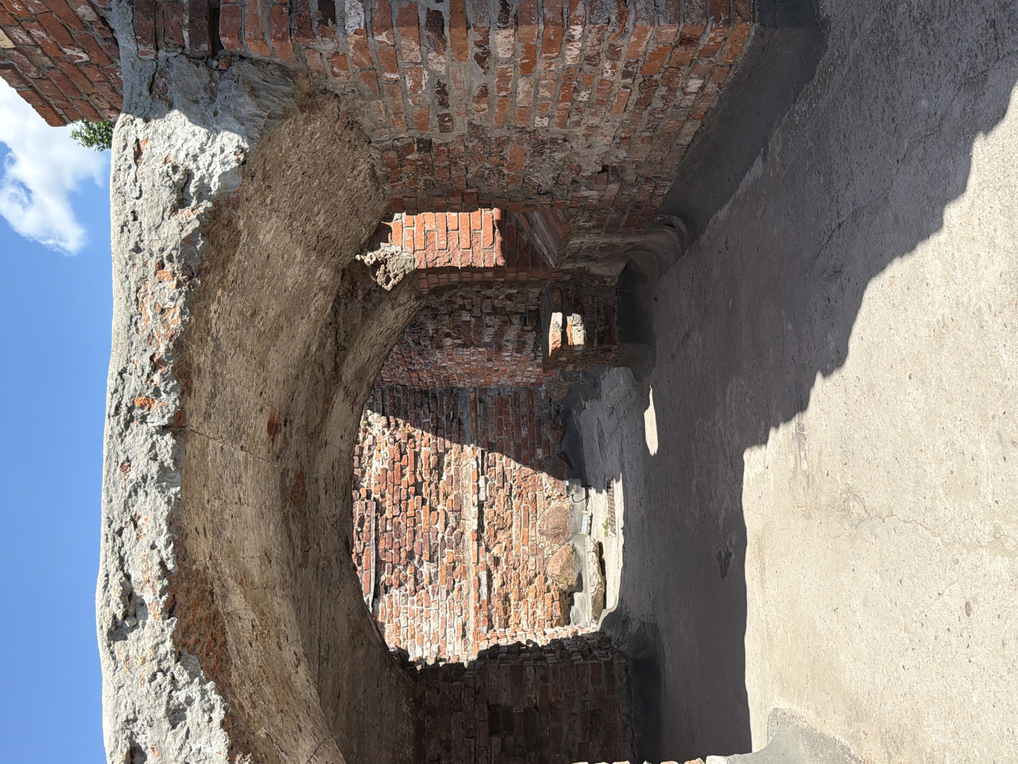An outdoor shot shows the inside of an arched stone structure, possibly a tunnel or walkway, made of brick. The arched ceiling appears to be constructed from stone and concrete with exposed brickwork. The walls are made of red brick, some of which appear aged and weathered. The ground is a light-colored concrete or stone surface, with shadows cast across it from the arched opening. In the background, more brick walls are visible, as well as large stones and the clear blue sky with a few white clouds. The scene suggests an old, possibly historical, site.