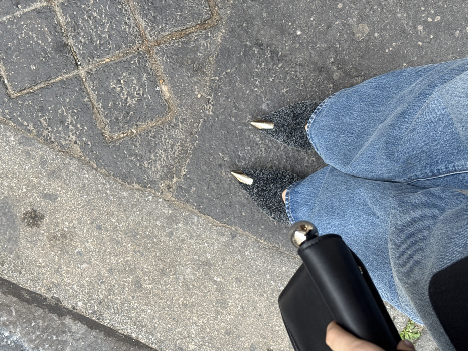 An overhead view captures the lower body of an individual standing on an outdoor pedestrian surface in Unknown, Unknown. The person wears relaxed-fit blue denim jeans and dark grey textured knit pointed-toe shoes, each featuring a prominent metallic gold-toned toe cap. The individual's left hand holds a black rectangular clutch or wallet, which has a spherical chrome-colored clasp or decorative element visible at its end. The ground beneath consists of two distinct paving materials: a lighter grey granular concrete or asphalt on the left side, diagonally intersecting with a darker, textured, square-patterned block paving on the right side.