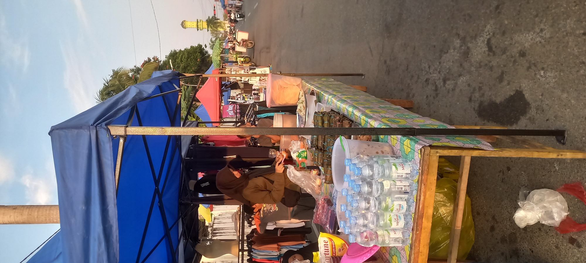 The image depicts a street market scene in Panji, Indonesia. A stall is set up with a blue canopy, displaying various items including clothing on racks and food products on a table. A woman in a hijab is seen interacting with the food items on the table. Bottles of water are neatly arranged on the table, along with wrapped food packages and a container of crackers. In the background, other stalls with colorful canopies are visible, as well as a minaret of a mosque. The street is paved, and there are motorcycles and people in the distance, suggesting a bustling market environment. The lighting indicates it is either early morning or late afternoon.
