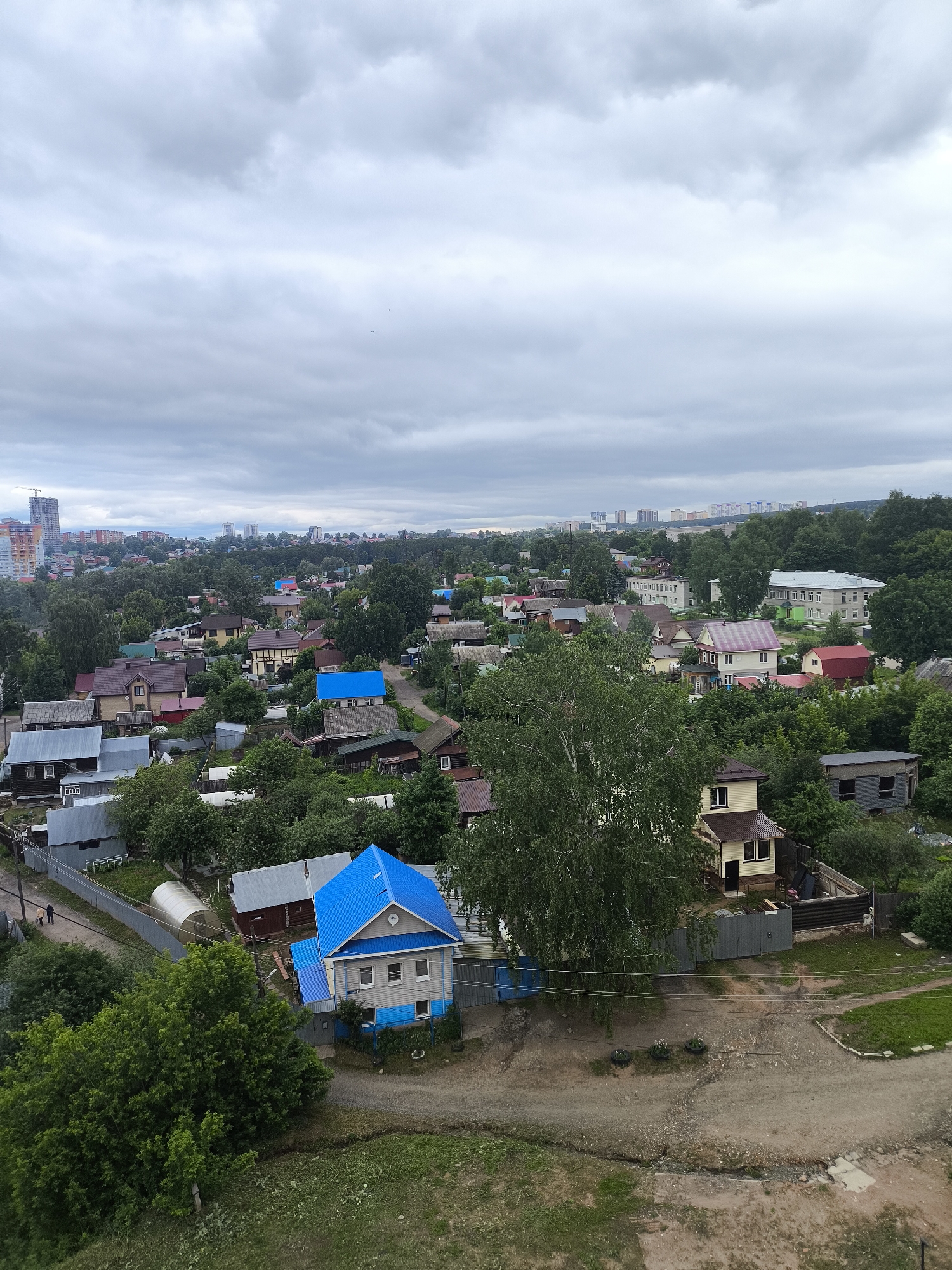 An aerial view presents a residential neighborhood under a uniformly overcast sky. In the foreground, a prominent two-story house features a bright blue pitched roof and light blue siding, with a smaller, similarly roofed extension. A large, leafy deciduous tree stands to its right. The ground in the immediate foreground is unpaved, composed of dirt and gravel, with interspersed patches of grass. Three tires are arranged as planters on the ground near the center. A narrow dirt path ascends towards the left, where two human figures, an adult and a child, are visible walking. Properties are delineated by various fences, including corrugated metal and wire mesh. A greenhouse structure is positioned behind a fence on the left.
The midground reveals a dense residential area comprising numerous single-family houses of varying architectural styles and roof colors, predominantly brown, red, grey, and some blue. These homes are heavily integrated with extensive green vegetation, including many trees and bushes, suggesting a well-treed environment. Unpaved roads and paths crisscross through this section. Further back in the midground, larger, multi-story buildings are observable, one featuring a distinct light green painted section.
The background is dominated by a cloudy, light grey sky. Below the horizon line, a distant cityscape is visible, characterized by multiple high-rise buildings. A tall construction crane stands prominently on the far left, indicating ongoing development.