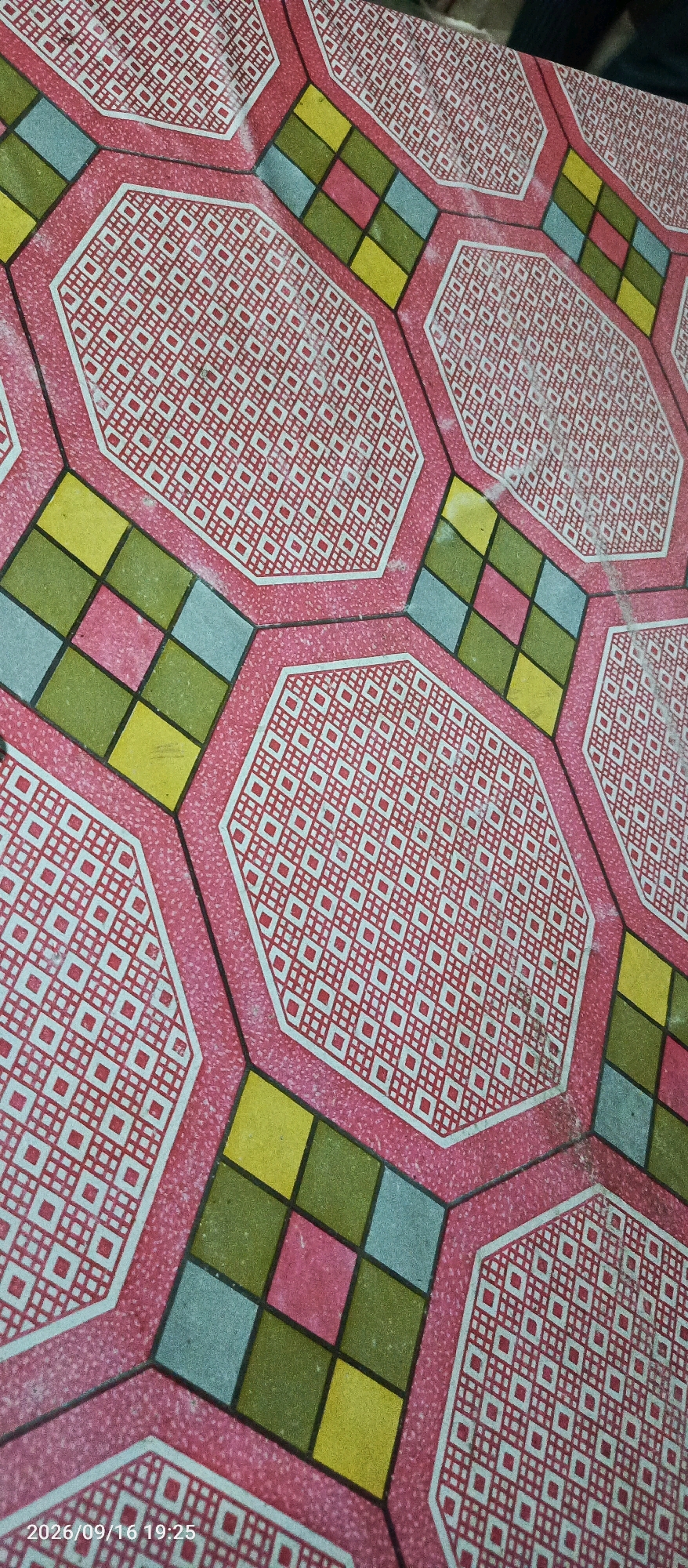 The image displays a close-up, high-angle view of a patterned floor. The floor features repeating hexagonal tiles with a pink border, each containing a white geometric pattern on a red background. Interspersed between the hexagons are smaller, square-shaped tiles in various colors: yellow, green, light blue, and pink. These colored squares are arranged in a diamond pattern within a larger hexagonal outline. The overall aesthetic suggests a tiled surface, potentially in an outdoor or indoor public space. The timestamp in the bottom left corner indicates "2026/09/16 19:25", and the text "Karangsono, Indonesia" is also visible. There are no people or animals present in the image. The lighting appears to be natural, casting subtle shadows and highlighting the texture of the floor.