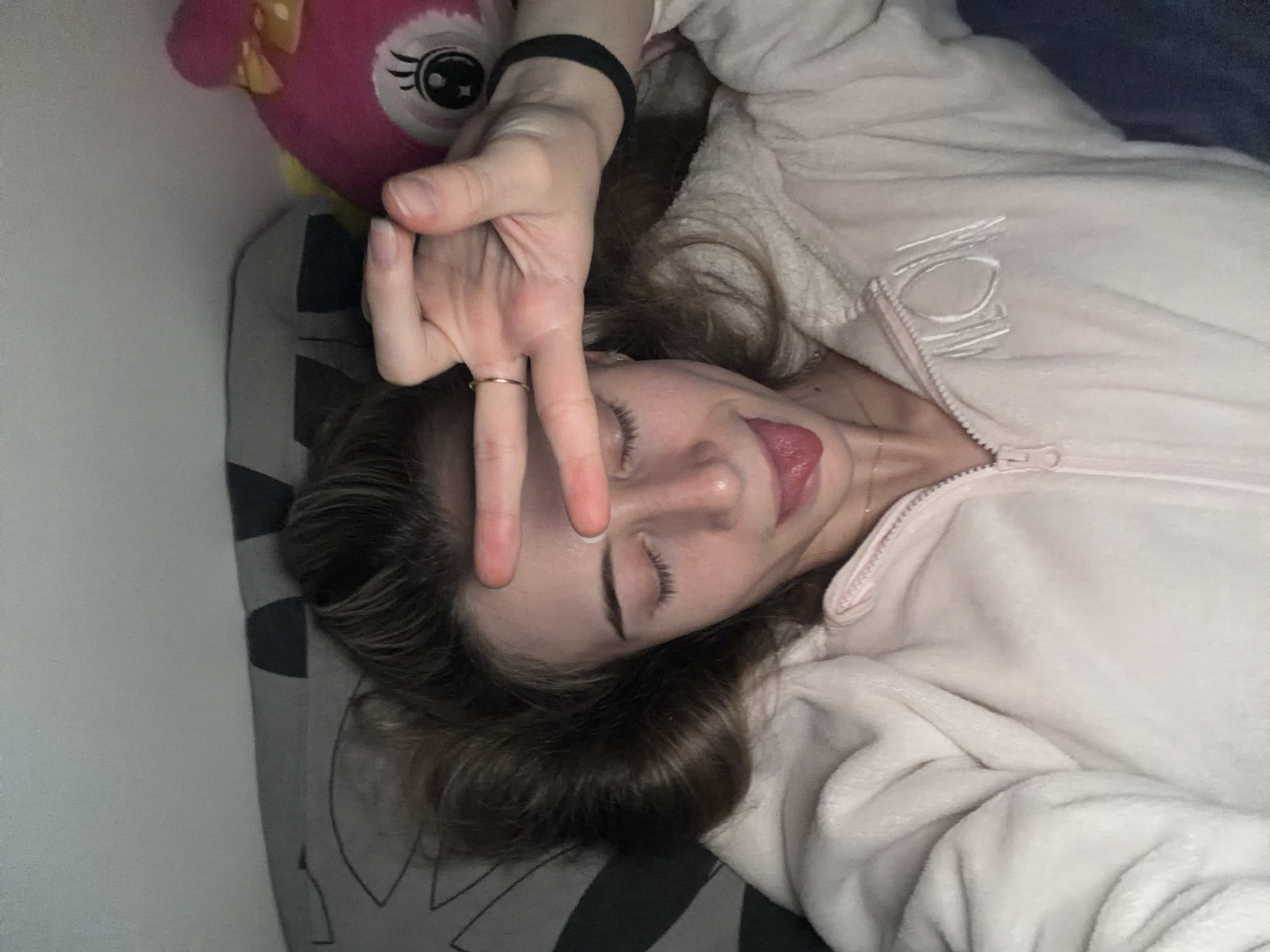 A young woman is lying on a pillow, possibly in a bed. She is wearing a light-colored, zip-up hooded sweatshirt, and a black hair tie is visible on her wrist. Her eyes are closed, and she is sticking out her tongue slightly. She is holding up two fingers in a peace sign, and a ring is visible on her finger. A pink, plush toy is next to her head on the left side. The pillow has a gray and black geometric pattern. There is no indication of Plovdiv, Bulgaria in the image.