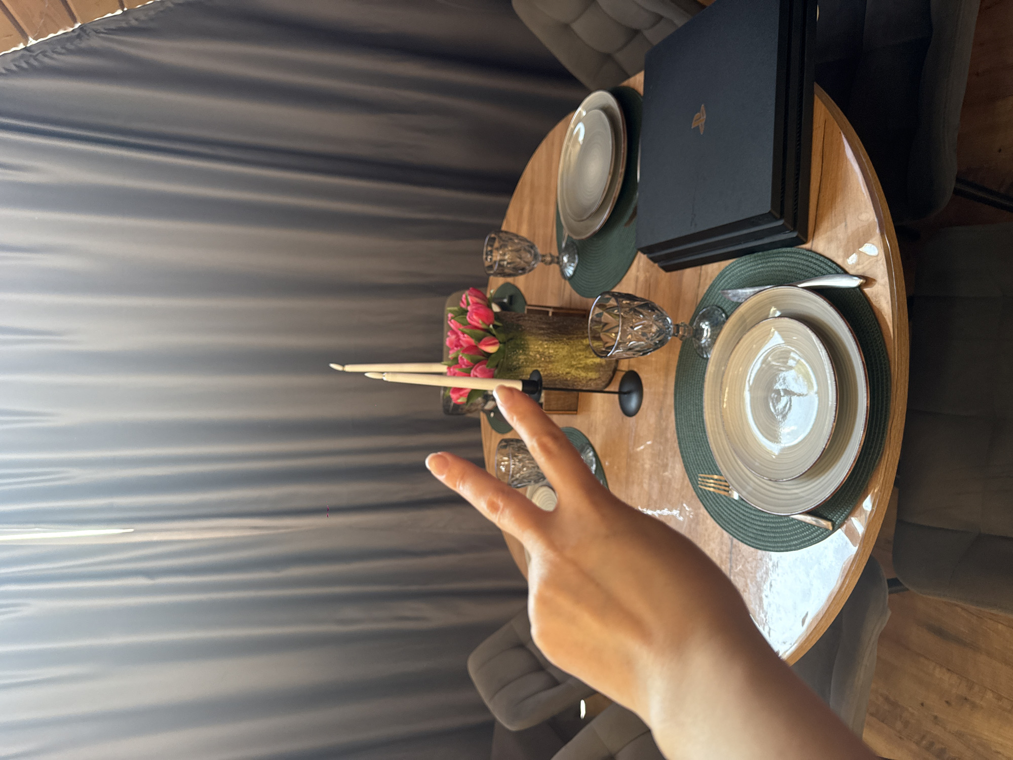 A round wooden dining table is set for two, with a floral arrangement and candles in the center, along with a PlayStation console. A hand making a peace sign is visible in the foreground. The room has grey curtains and modern chairs. Location information is not discernible from the image.