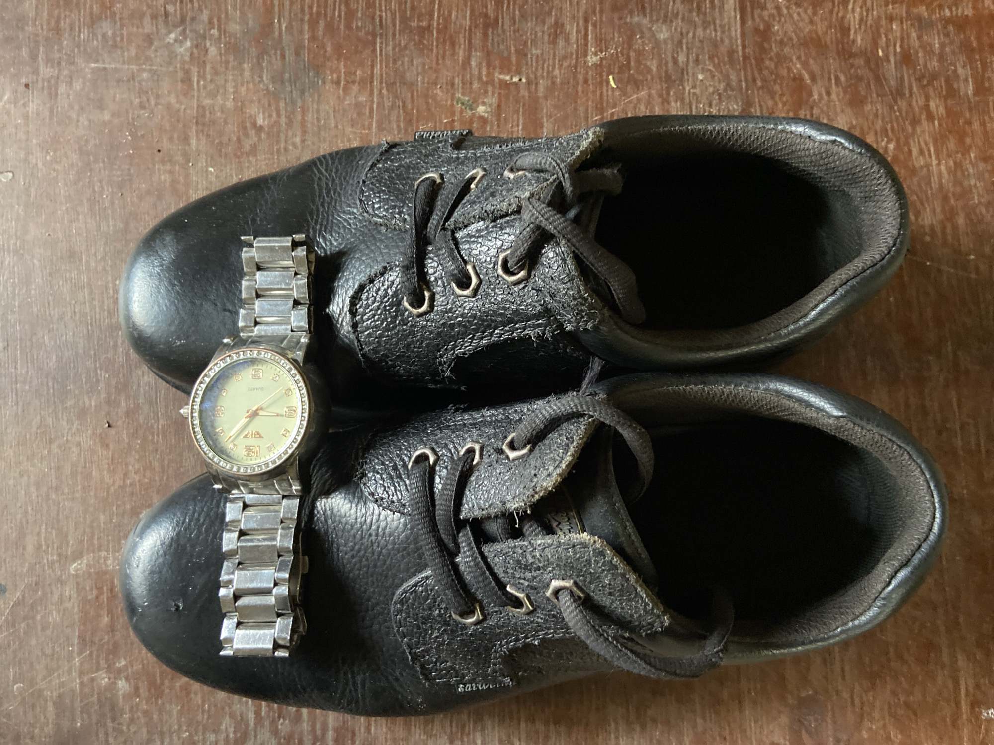A pair of black leather work boots with laces is shown from a top-down perspective, placed on a wooden surface. A silver-banded wristwatch with a light-colored face and diamond accents is resting on the top of the boots, bridging them. The setting is an indoor or outdoor wooden surface, possibly a floor or table, with a grainy texture. The image appears to be taken in Mararaba, Nigeria. There are no people or active interactions depicted.