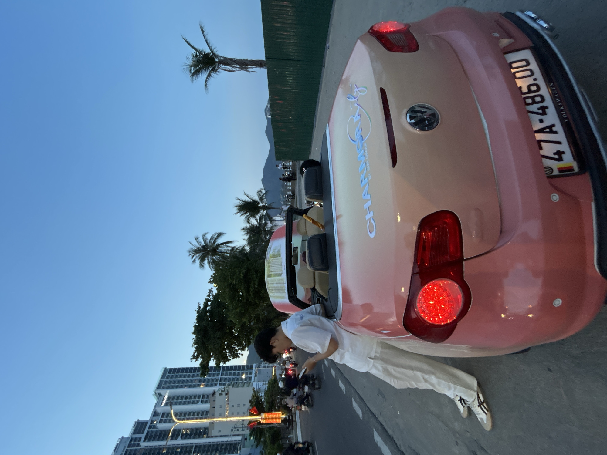 A wide-angle, upward-tilted shot captures a partially visible pink Volkswagen New Beetle convertible parked on the side of a street in Bắc Nha Trang, Vietnam. A person dressed in all white stands near the open car door, appearing to be looking at a phone. The car's license plate reads "47A-486.00". Buildings with modern architecture line the street, interspersed with tropical trees. Streetlights are visible. The sky is clear and blue.