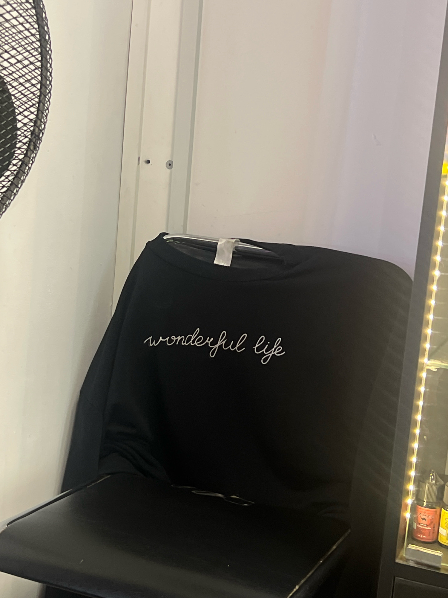 A black sweatshirt with "wonderful life" embroidered in white cursive script is draped over a black chair. The sweatshirt is hanging on a hanger. To the left of the sweatshirt, part of a black fan is visible against a white wall. To the right of the sweatshirt, a black cabinet with several small bottles and yellow LED lights is visible. The scene appears to be indoors.