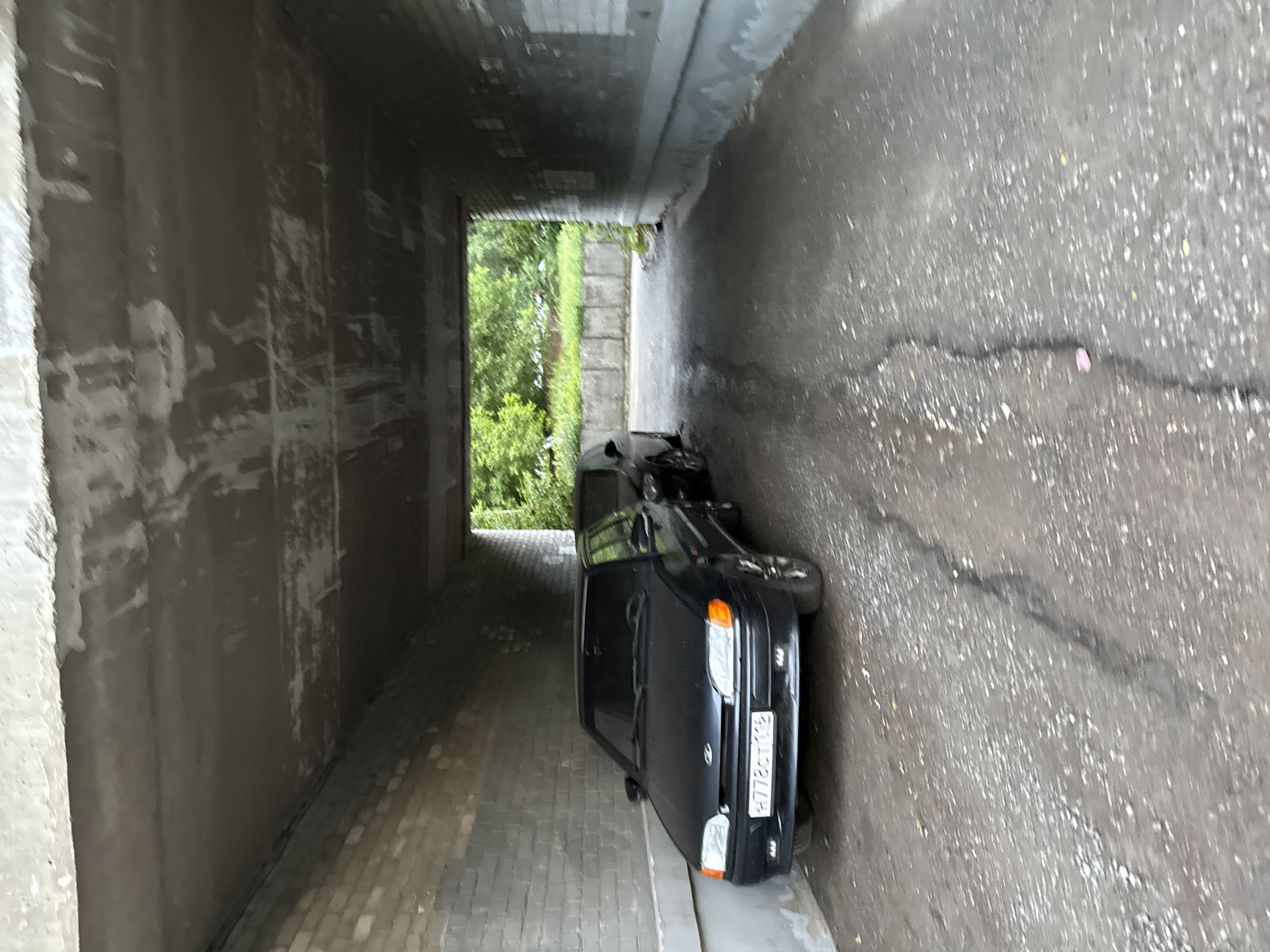 An underpass in Kazan, Russia, has two black cars parked inside. The first car, a sedan, is positioned on the left, facing forward. It has a white license plate with Russian characters and numbers that read "H778CT 116". The second car, also a sedan, is parked behind the first and is partially obscured. The underpass has a rough, concrete ceiling and brick walls. The ground is a mix of gravel and asphalt, with a visible crack in the center. Outside the underpass, there is greenery and a concrete barrier.