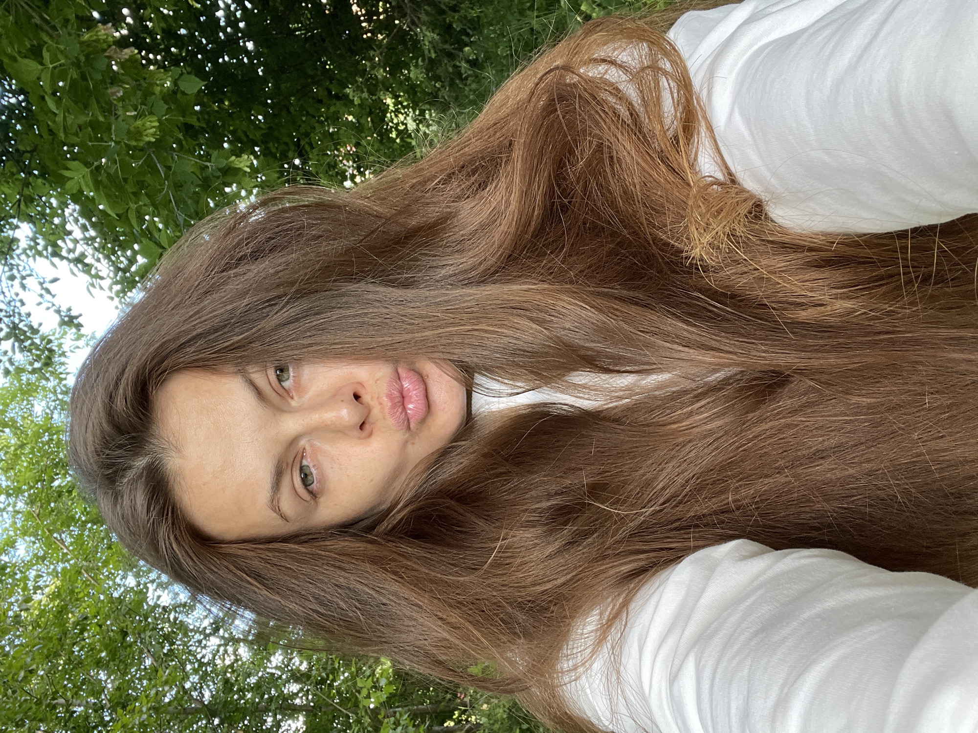 A medium shot shows a woman with long, brown hair and green eyes looking at the camera and puckering her lips. She is wearing a white top. The background is blurry green foliage, suggesting an outdoor setting. No specific landmarks in Заречный, Russia, are identifiable.