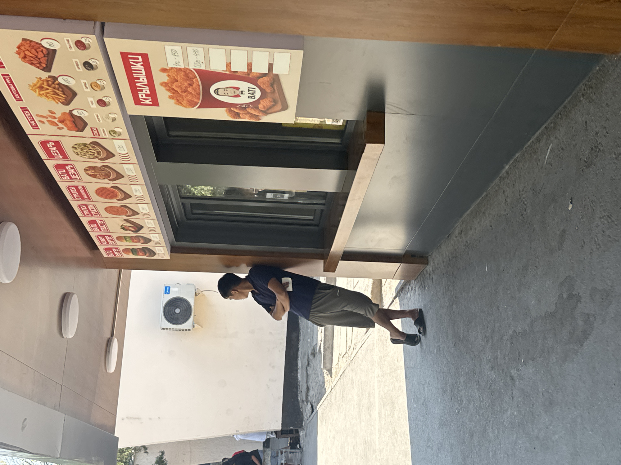 The image shows a young man in a dark blue polo shirt and grey shorts, leaning against a pillar next to a fast-food window. He is looking to his right, with his arms crossed. The fast-food establishment has a menu board displaying various food items, including chicken wings, fries, and burgers, with prices in rubles. The outdoor setting appears to be sunny, with a concrete ground and a light-colored wall in the background. The overall impression is that of a casual encounter at a food vendor. The presence of Russian currency and Cyrillic script on the menu suggests the location is likely in a region where Russian is spoken, such as Bishkek, Kyrgyzstan, a possibility supported by the presence of an air conditioning unit on the wall, common in warmer climates.