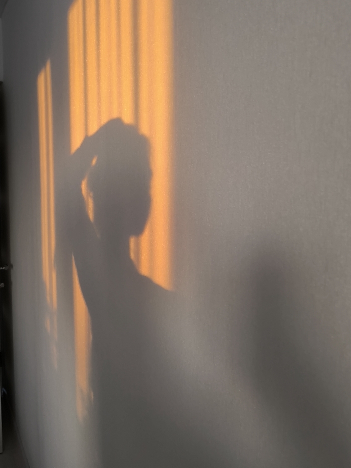 A stark white wall bathed in the warm, golden light of sunset showcases striking shadows. Vertical beams of light create textured patterns, interspersed with the dark silhouette of a person. The figure raises a hand to their head, casting a larger, diffuse shadow alongside their body. The interplay of light and shadow conveys a sense of intimacy and quiet contemplation.