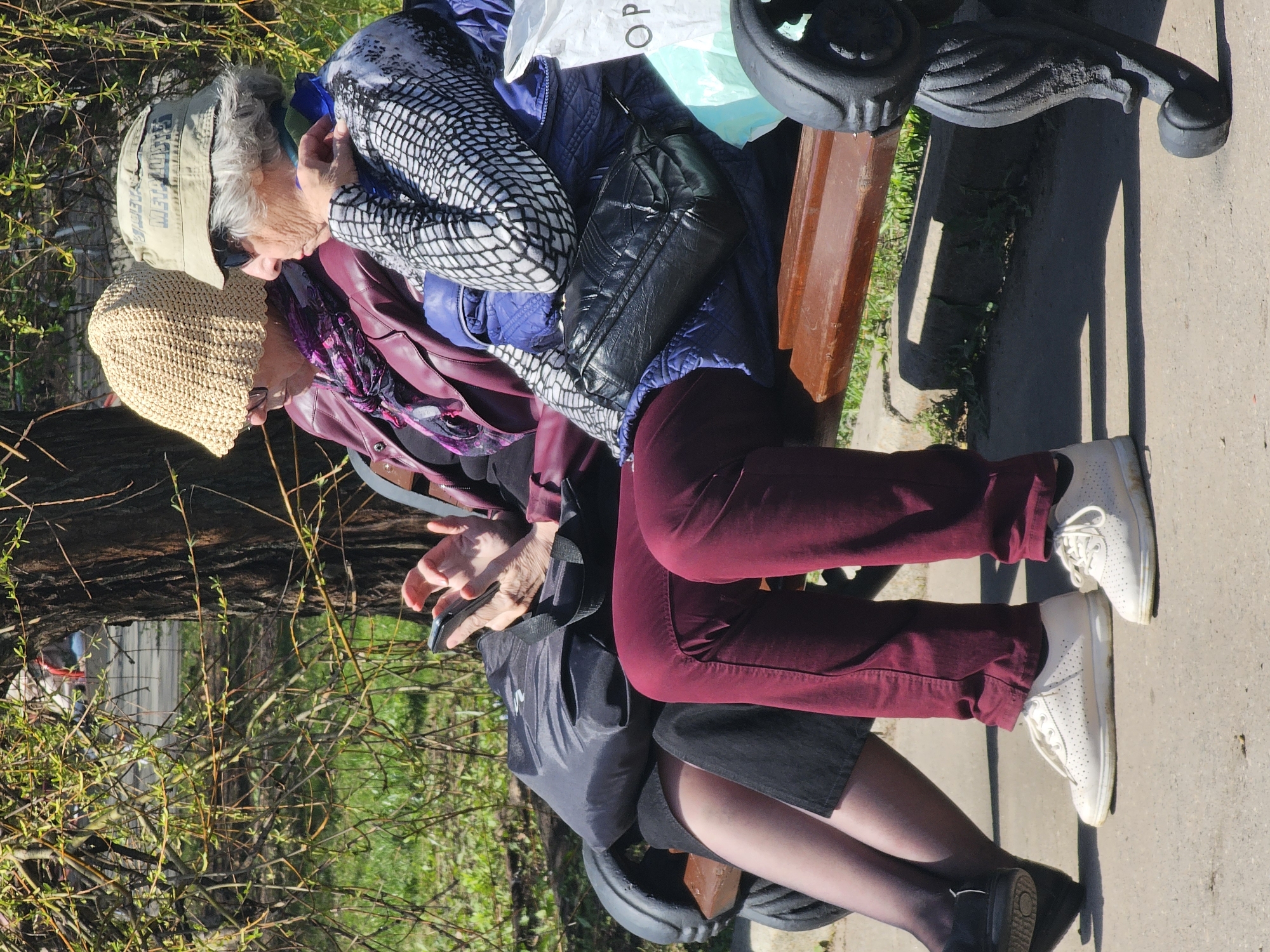 Two older women sit on a park bench in what appears to be Moscow, Russia. The woman on the right wears a baseball cap, a black-and-white patterned jacket, a blue jacket, and maroon pants with white sneakers. She appears to be speaking to the other woman. The woman on the left wears a straw hat, a purple jacket, and a floral scarf. She wears a black skirt, black stockings, and black shoes. The bench is dark brown wood with black wrought iron supports. A black purse and shopping bag sit on the bench between the women. Trees and bushes with budding leaves are visible in the background. A sidewalk is adjacent to the bench.