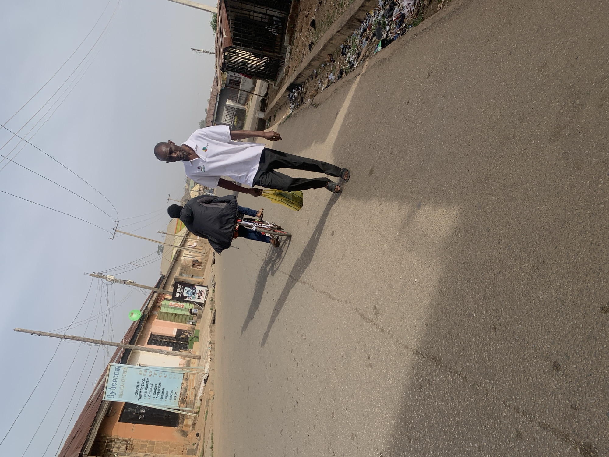 A man in a white shirt and dark pants walks down a paved road carrying a yellow bag. Next to him, another person is riding a bicycle. The street is lined with buildings, some of which appear to be businesses. The Cybercom Business Centre sign is visible. The scene is in Jos, Nigeria. There is trash along the side of the road.