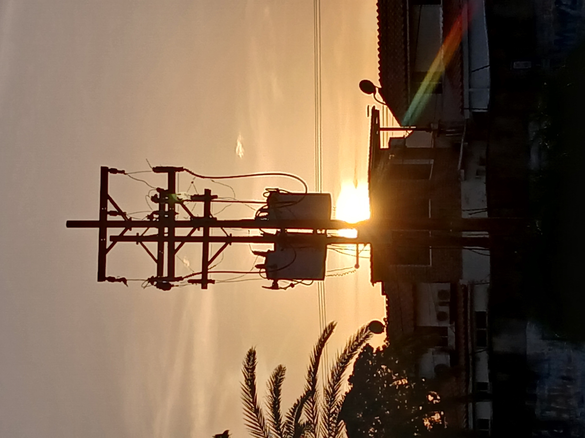 The image shows a cityscape at sunset, likely in Ciudad Guayana, Venezuela, given the presence of palm trees and local architecture. An electrical pole with transformers and wiring is silhouetted against the bright sun. A two-story building with a red-tiled roof and a satellite dish is partially visible to the right. Vegetation is visible at the bottom. The overall scene depicts an urban environment during twilight.
