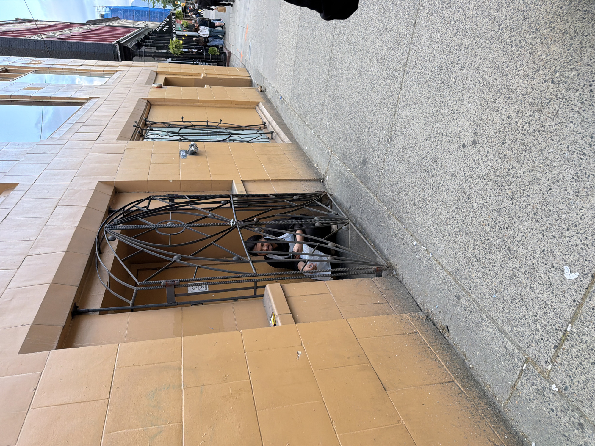 The image depicts a street scene in Vancouver, Canada, with a person visible behind a wrought iron grate covering a basement window of a building with a tan tiled facade. The person appears to be interacting with something inside the building, possibly handling white plastic bags. The sidewalk is concrete and runs adjacent to the building. Other windows with wrought iron details are visible higher up on the building. In the background, other buildings and pedestrians can be seen, including signage for "Danny's Inn" and "Coffee Rooms." A blue construction tarp is visible covering the upper part of a building in the distance.