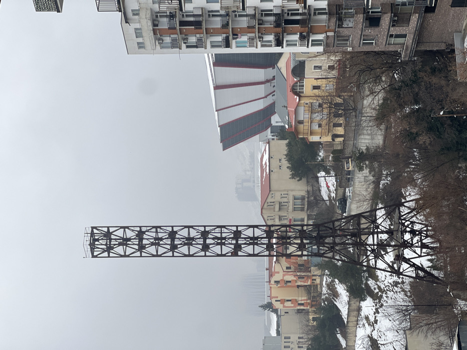 An upward-angled view reveals a cityscape under an overcast sky. In the foreground, a long, dark metal structure with a grid-like pattern extends horizontally. Beyond it, buildings of varying heights and architectural styles fill the frame, including a striking white structure with red accents. Bare trees line a road or walkway, adding depth to the scene. The overall impression is urban, with a sense of coldness due to the gray sky and dormant vegetation.