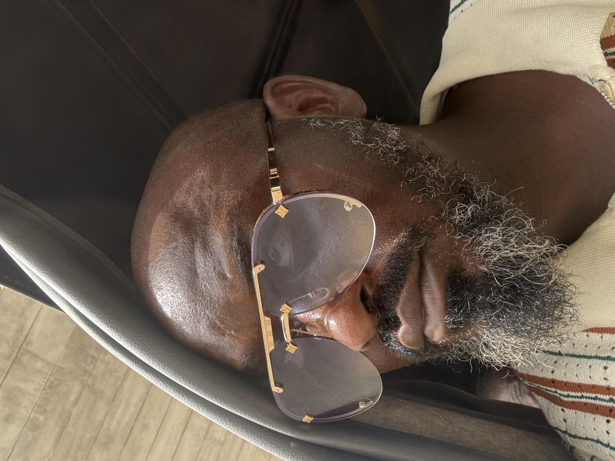 A close-up shot depicts a bald, middle-aged Black man with a graying beard asleep in what appears to be a black leather chair. The man's face is positioned sideways, obscuring most of his features except for his nose, mouth, and ear. A pair of sunglasses rests on his face, obscuring his eyes. He is wearing a light-colored sweater with brown and green stripes. The background includes the leather chair and the floor. The location is not evident from the image. It is impossible to confirm that the picture was taken in Lekki, Nigeria, or anywhere else.