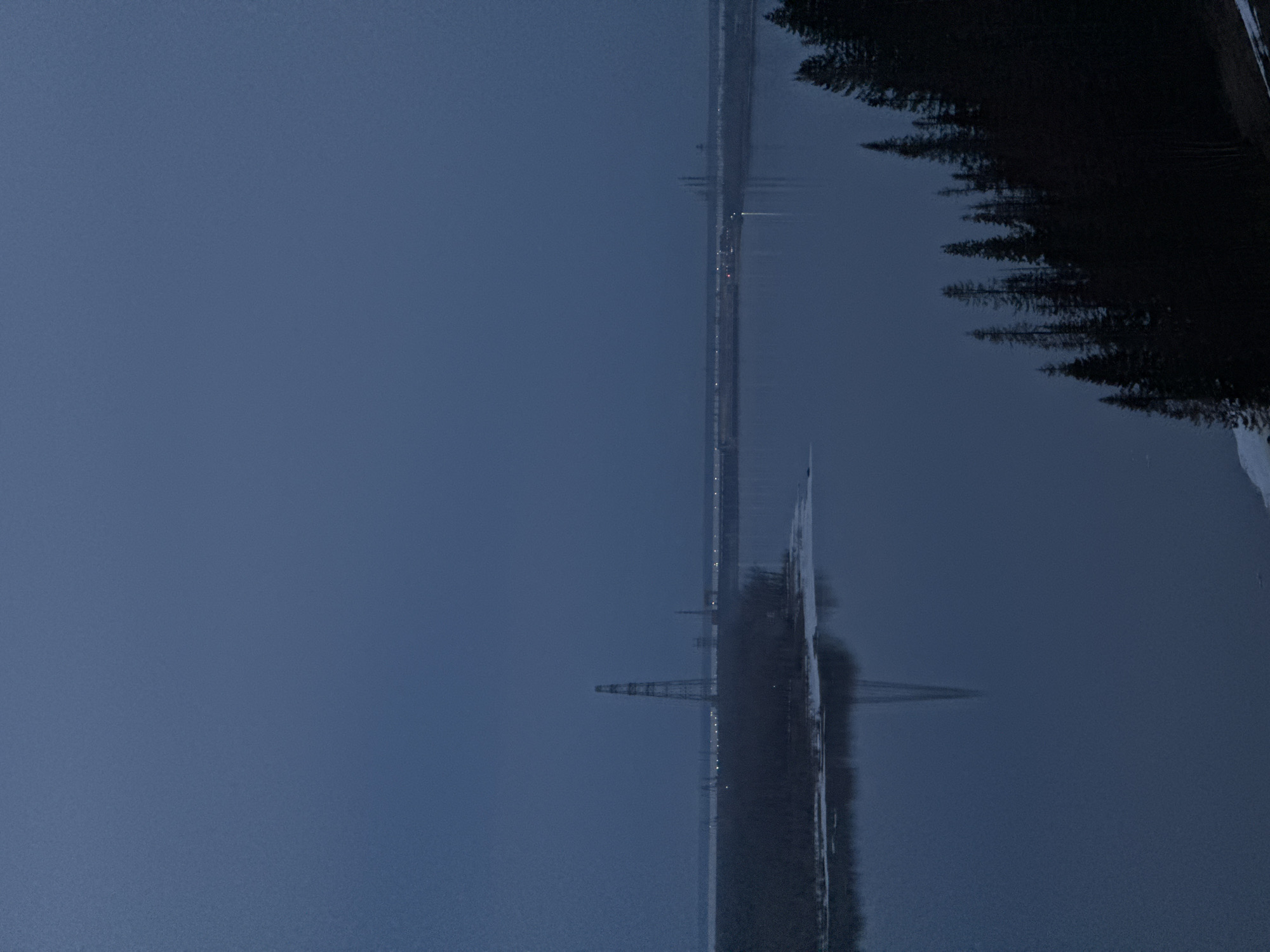 A body of water reflecting a foggy, overcast scene. A bridge with lights crosses the water horizontally across the image. The silhouette of dark trees lines the right edge of the image. An electrical tower can be seen on the left side. The location is Ольховка, Russia.