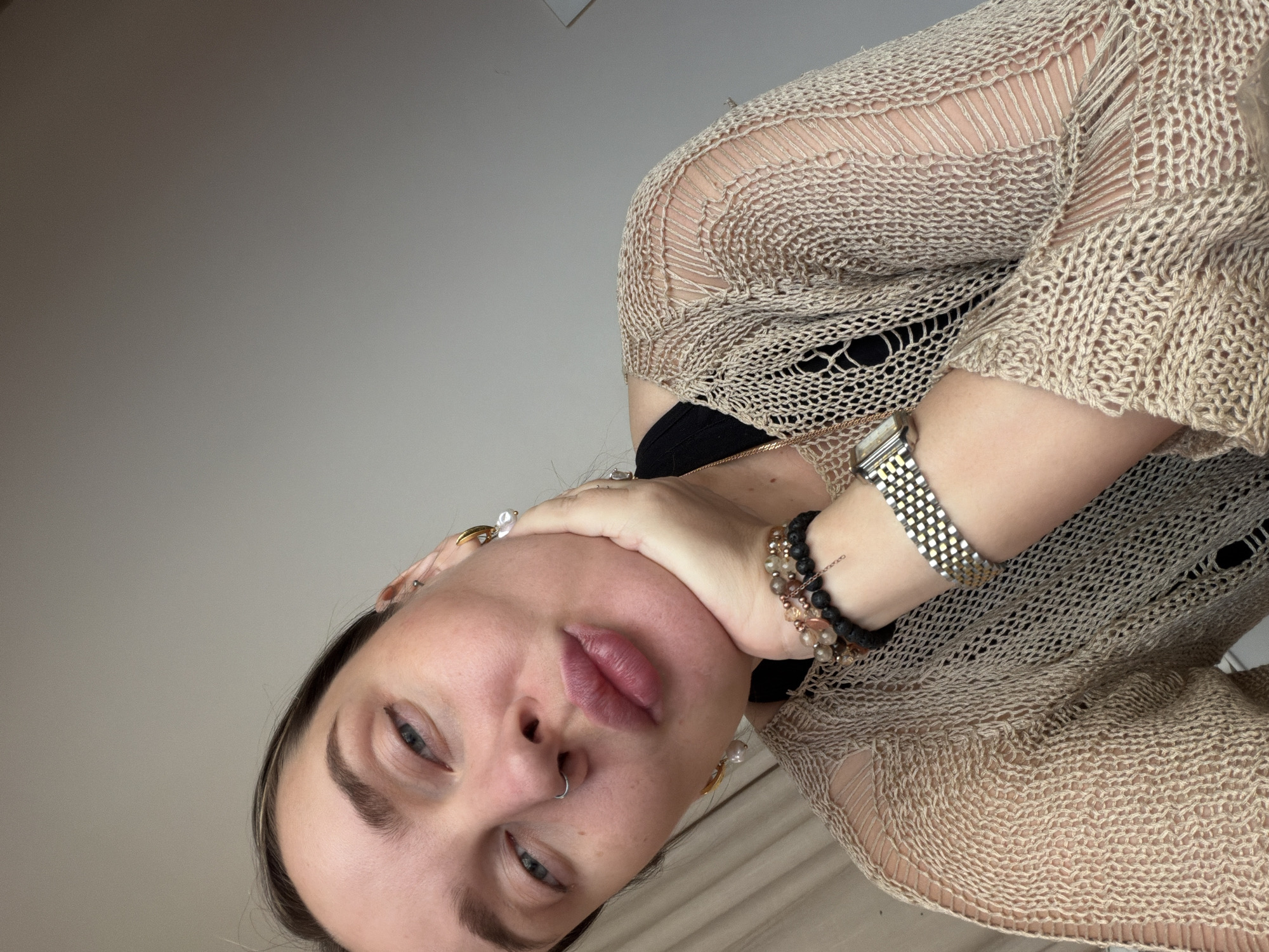 A woman with fair skin, light blue eyes, dark eyebrows, and a nose piercing is looking at the camera. She is wearing a beige, loosely knit sweater over a black top. She has a silver watch and a beaded bracelet on her wrist. Her hand is partially covering her face. The background is a plain, light-colored wall and a glimpse of what appears to be bedding. The image appears to be taken indoors. The location is Unknown, Unknown.