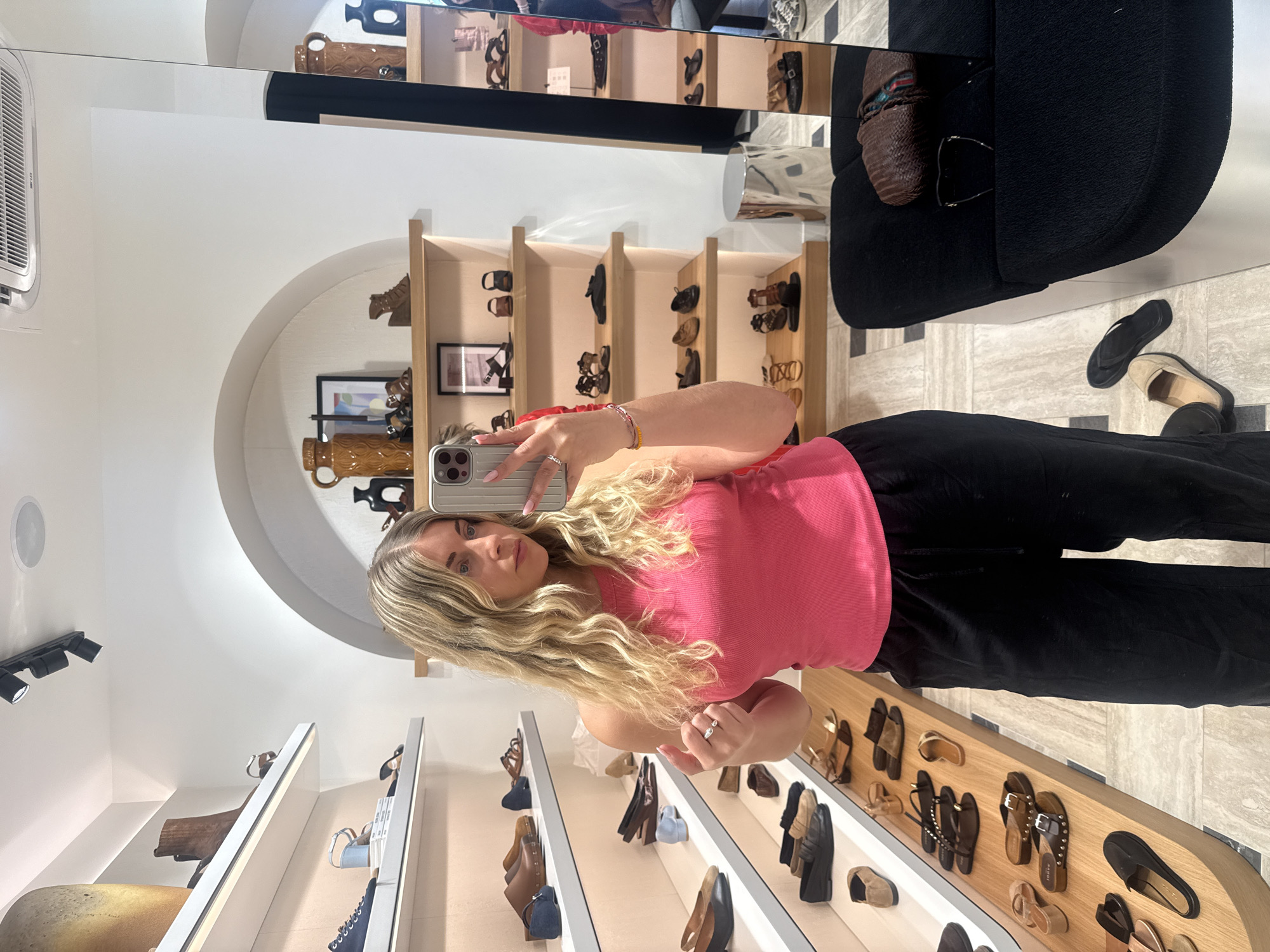 A blonde woman takes a selfie in a shoe store in Paris, France. She wears a pink tank top and black pants. Various styles of footwear are displayed on shelves and a table. A black chair is nearby.