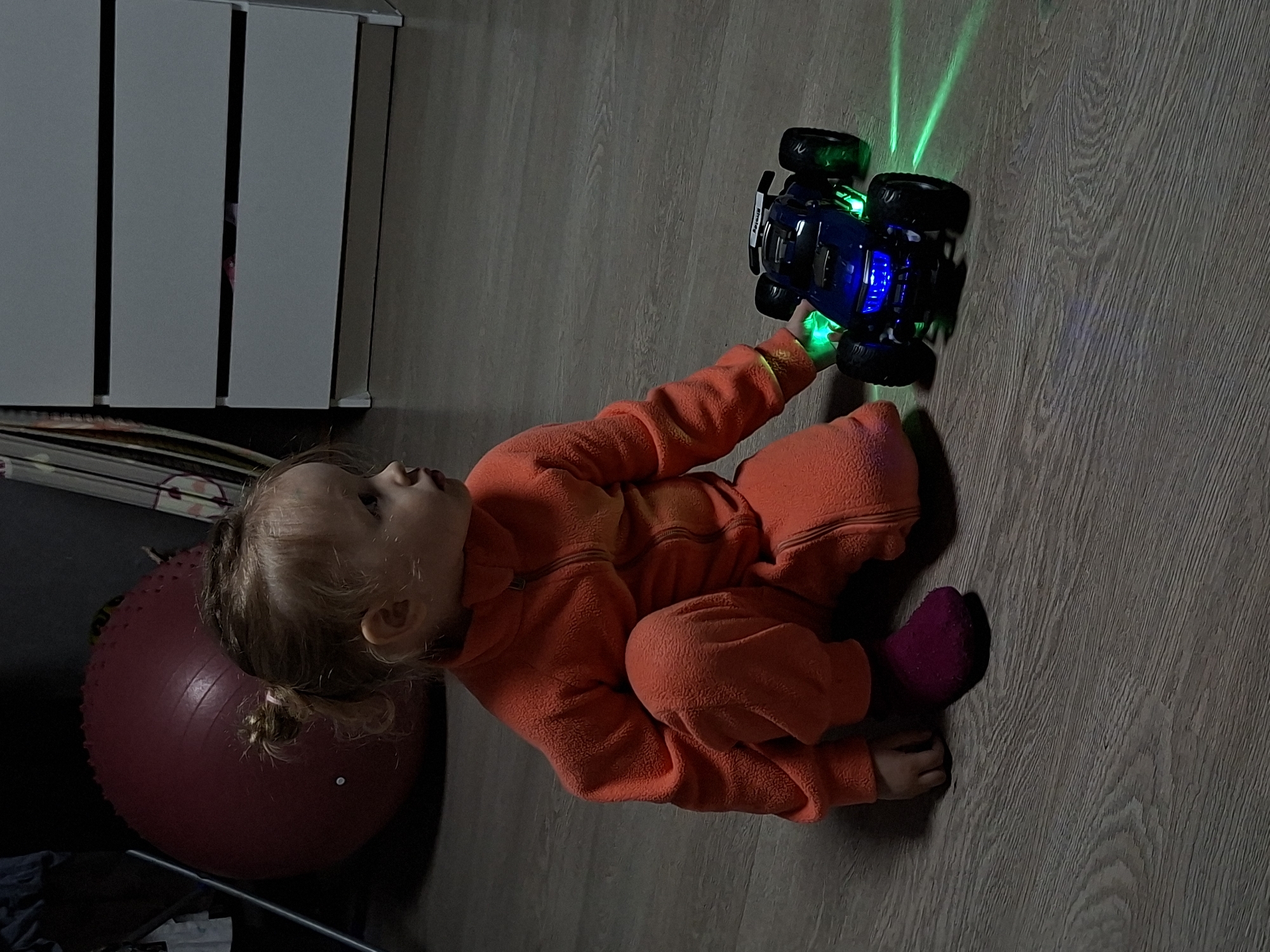 A young child sits on a gray wooden floor, wearing an orange fleece pajama set and one pink sock. The child is holding a blue toy truck that emits green and blue light beams. Adjacent to the child is a large, dark red exercise ball. In the background, a light-colored, vertically paneled storage unit is visible.